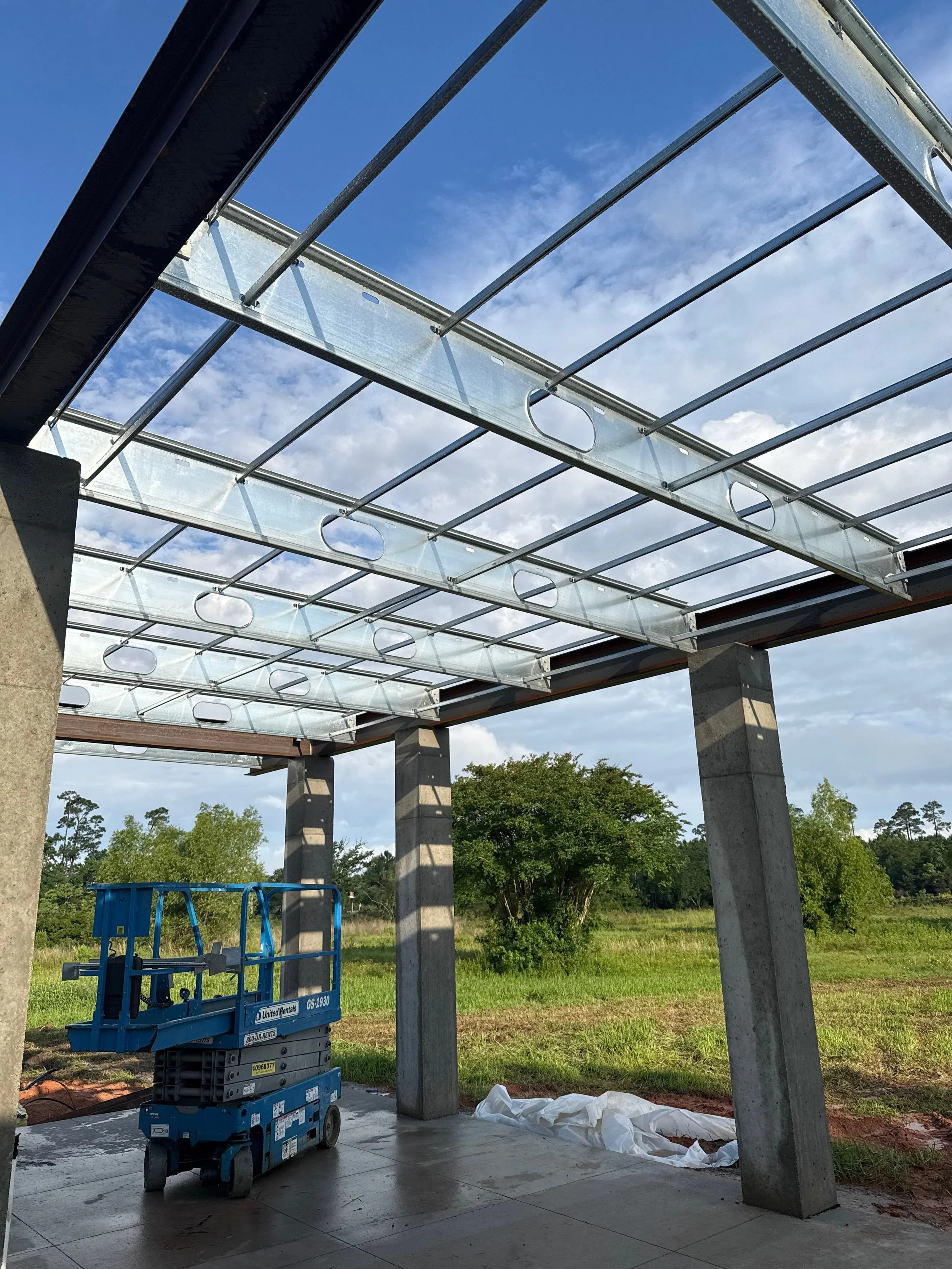 Construction site showing a steel framework for a roof with concrete columns, a blue lift, and a grassy area with trees in the background.