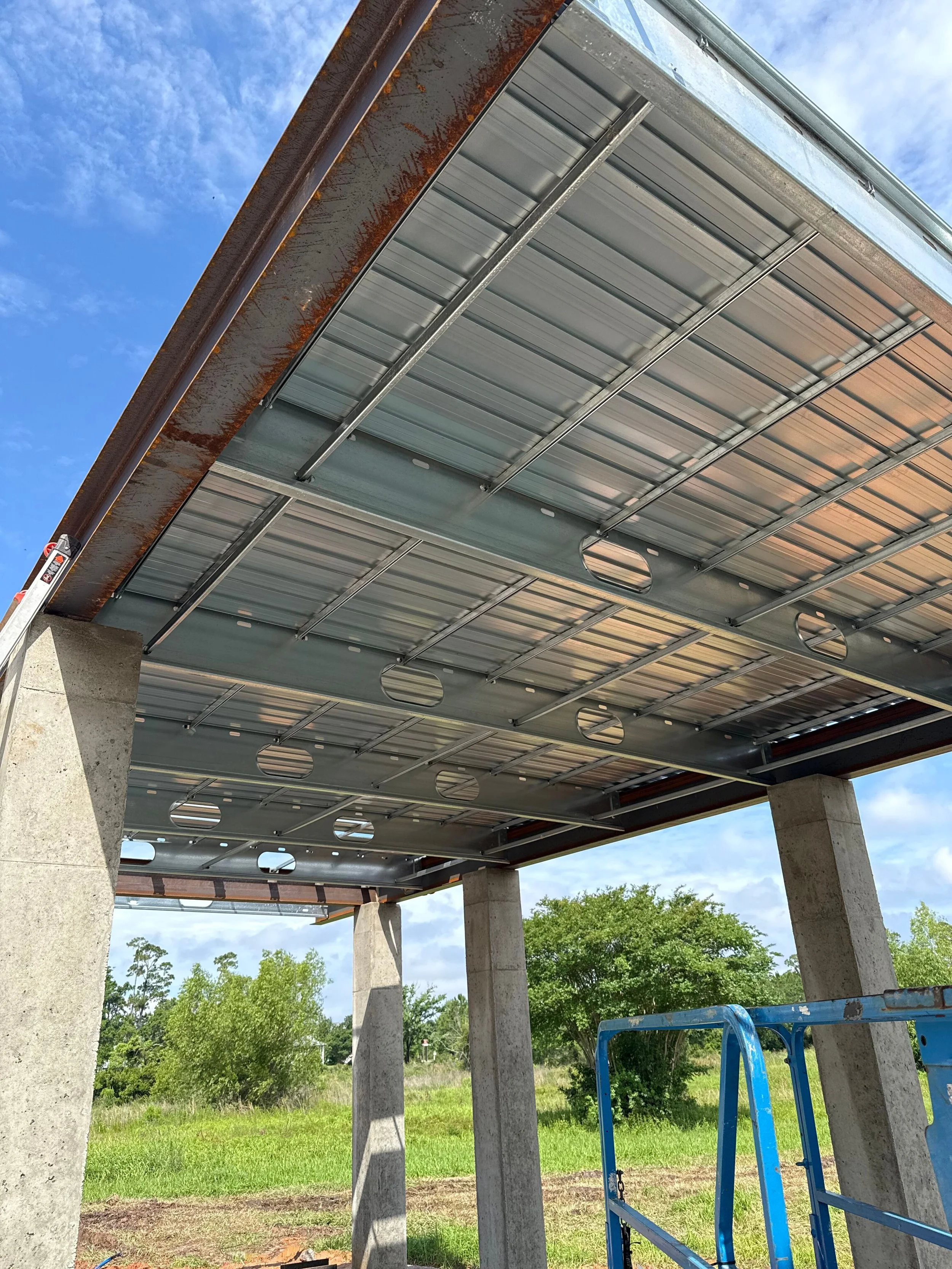 Construction site showing a concrete structure with steel support beams and a metal roof, supported by concrete pillars, with trees and blue sky in the background.