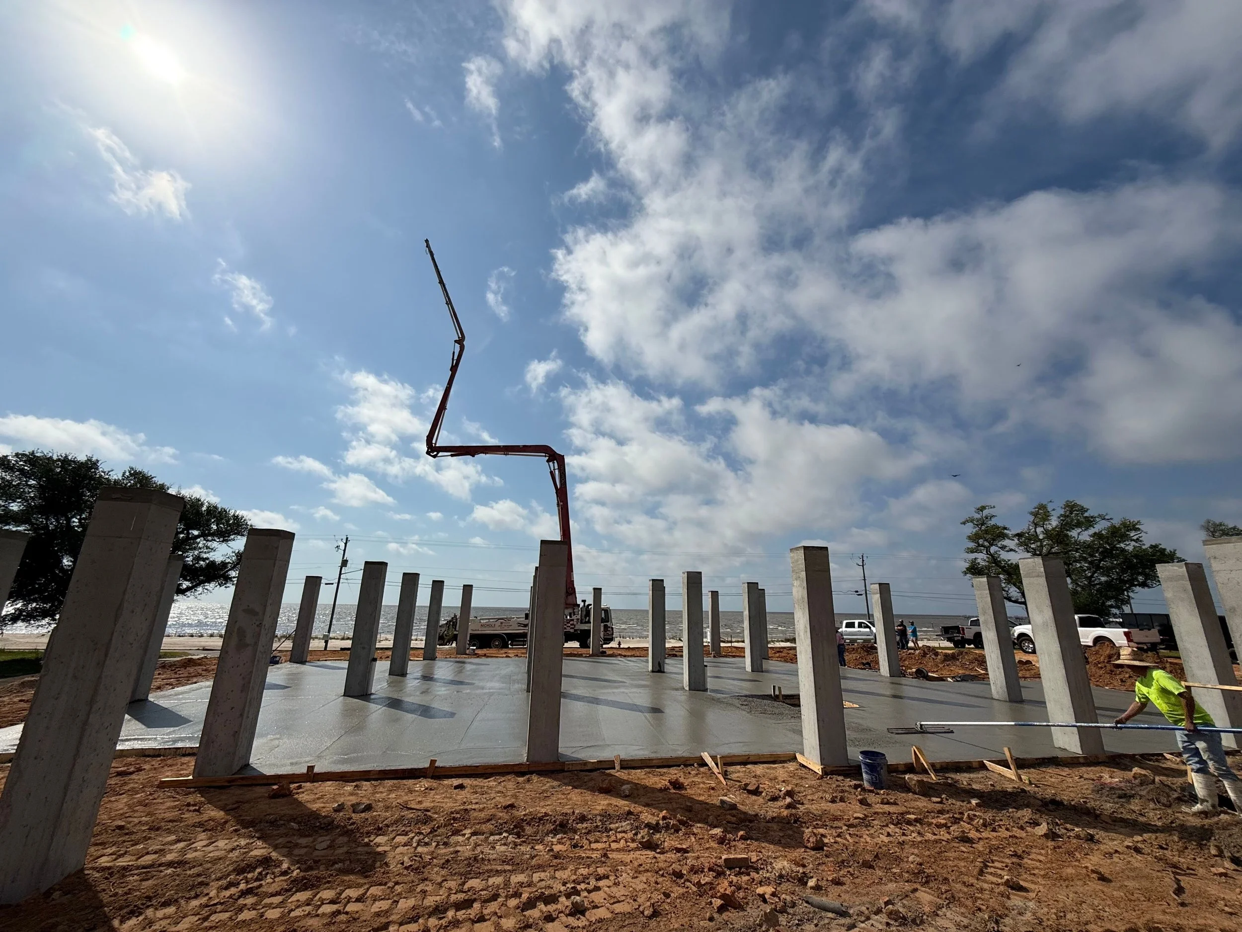 Construction workers building a structure with concrete columns, with an orange boom crane and partly cloudy sky in the background.