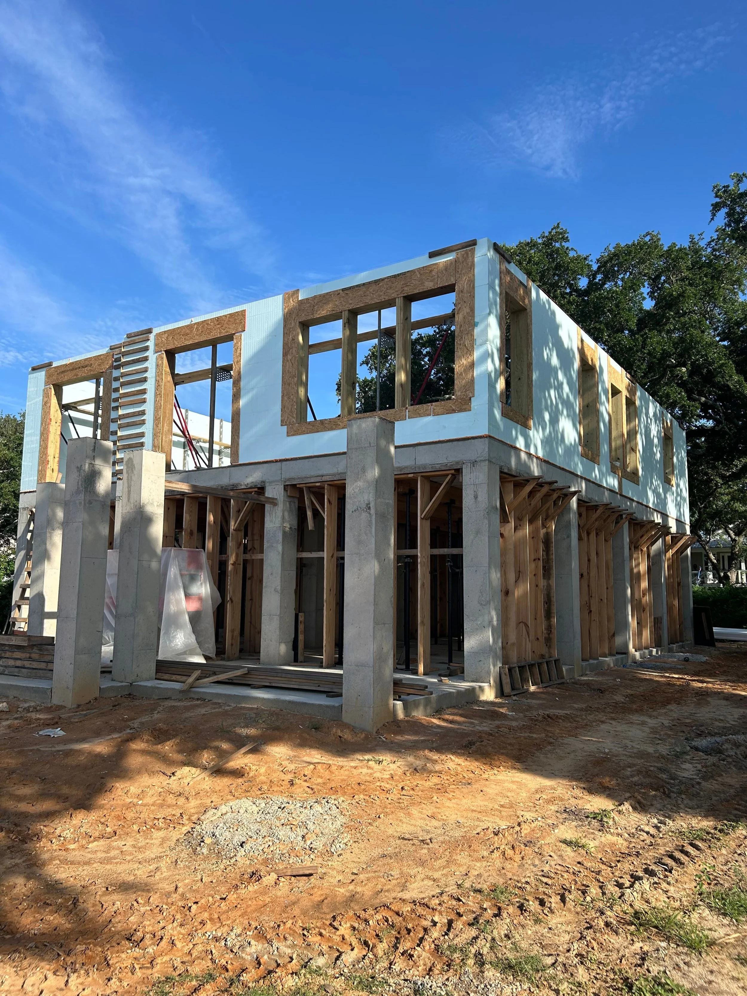 Construction site of a two-story building with wooden framing, concrete pillars, and some insulation panels, under a blue sky with scattered clouds.