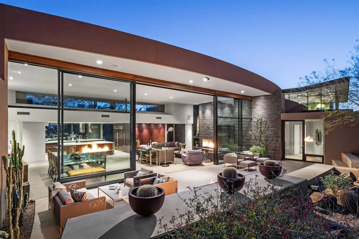 Modern house with large glass windows and patio, furnished with outdoor seating, potted plants, and desert cacti, under a clear evening sky.