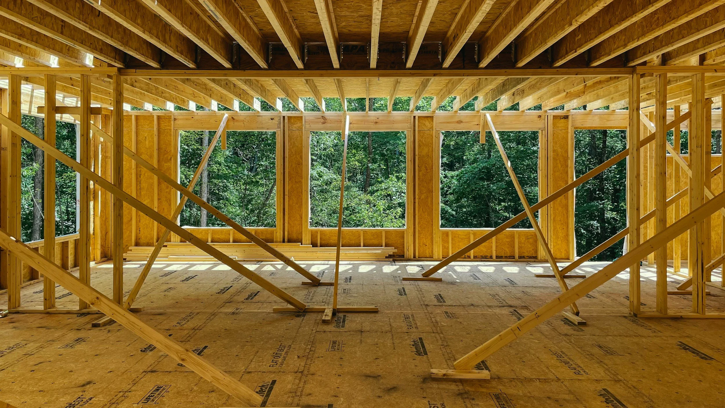 View of the wooden framework of a house under construction with exposed roof beams and wall studs, surrounded by a lush green forest outside.