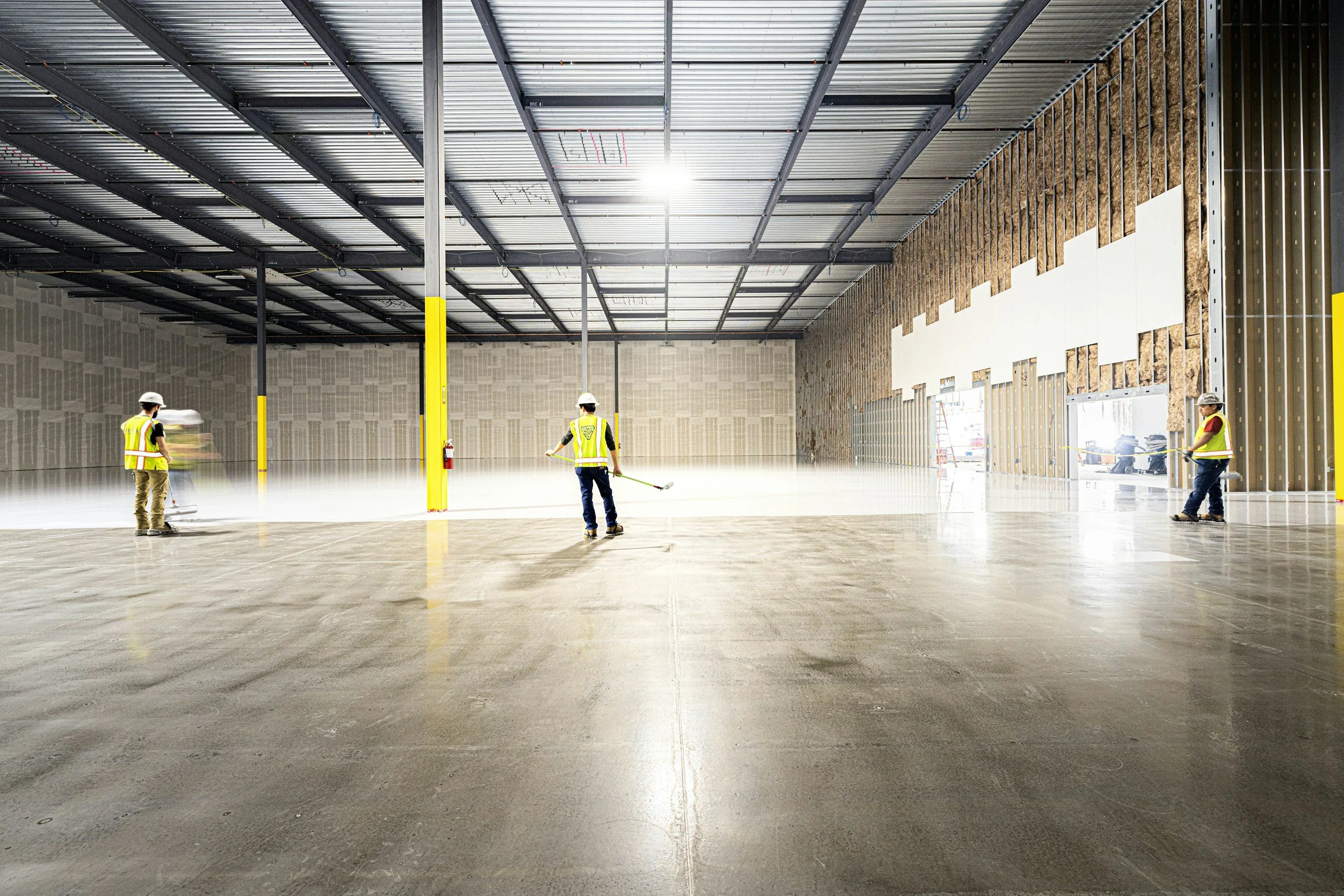 Construction workers in safety vests and hard hats working inside a large, unfinished building with exposed insulation and metal framework.