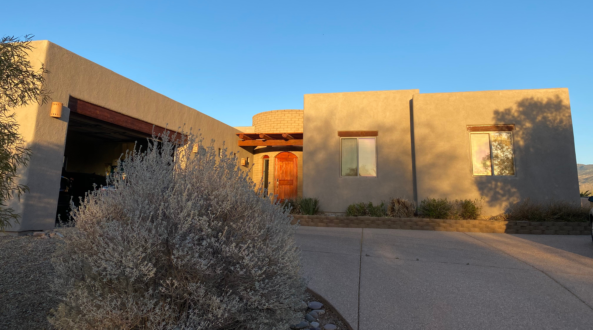 A modern house with a flat roof and stucco exterior, surrounded by desert landscaping, with the setting sun casting warm light on the facade and clear blue sky in the background.