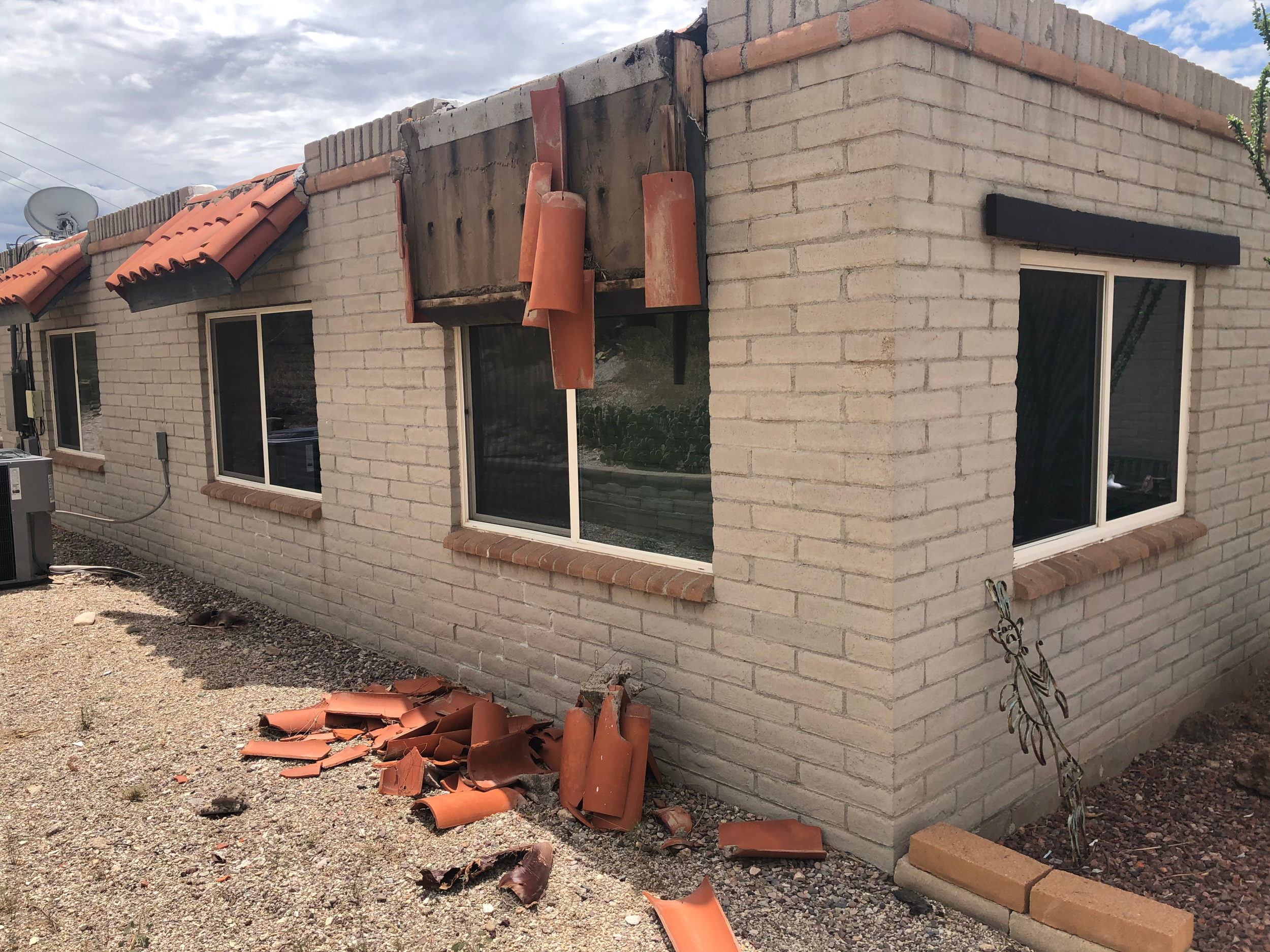 Side view of a house under construction with exposed brick walls, some roof tiles removed, and construction debris on the ground, including roof tiles and bricks.