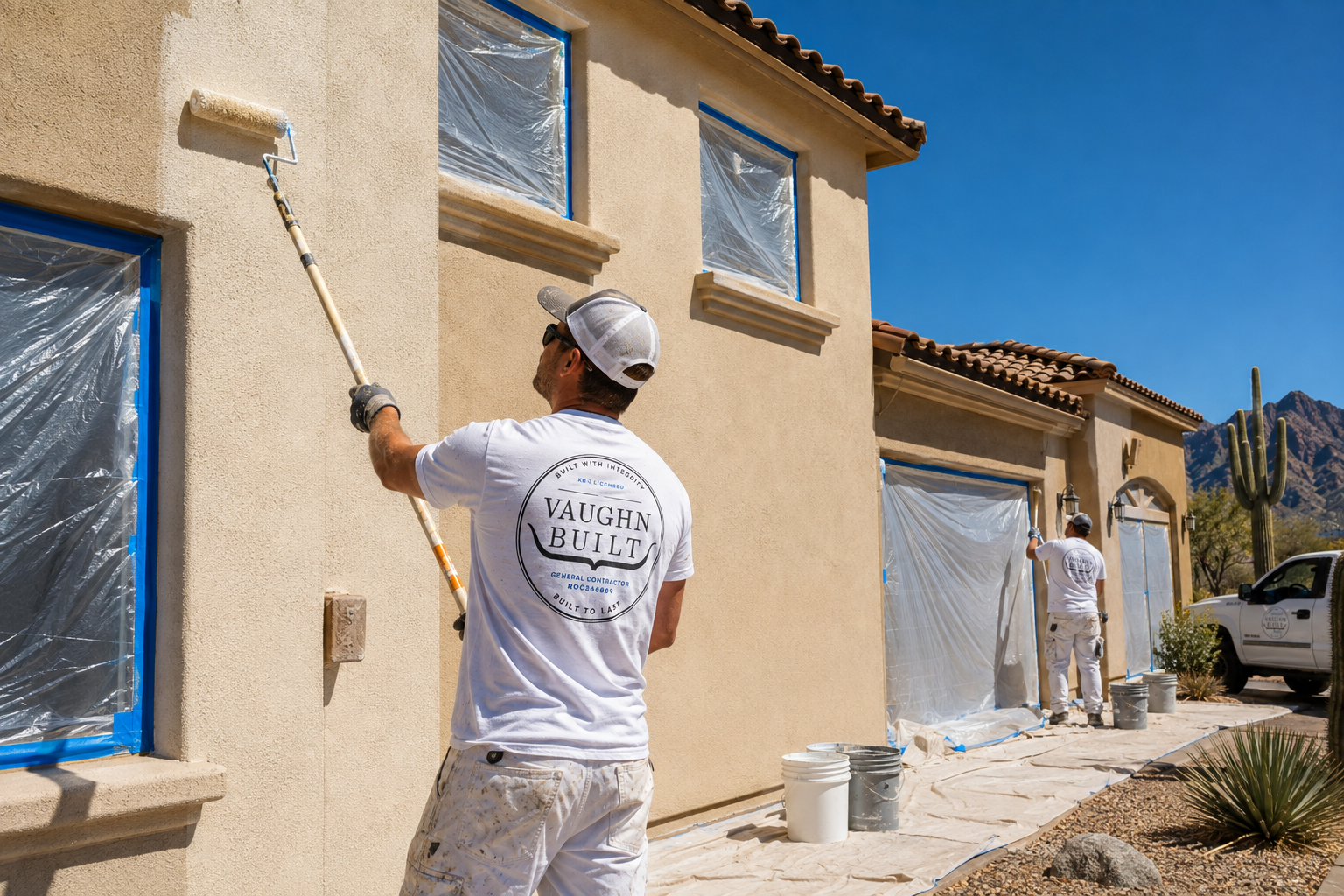 Two painters applying a fresh coat of paint to a house exterior with a paint roller. The house has protective plastic covering the windows, and the workers are dressed in white paint suits with gloves and caps. The scene is set in a sunny, arid landscape with mountains and a cactus in the background.