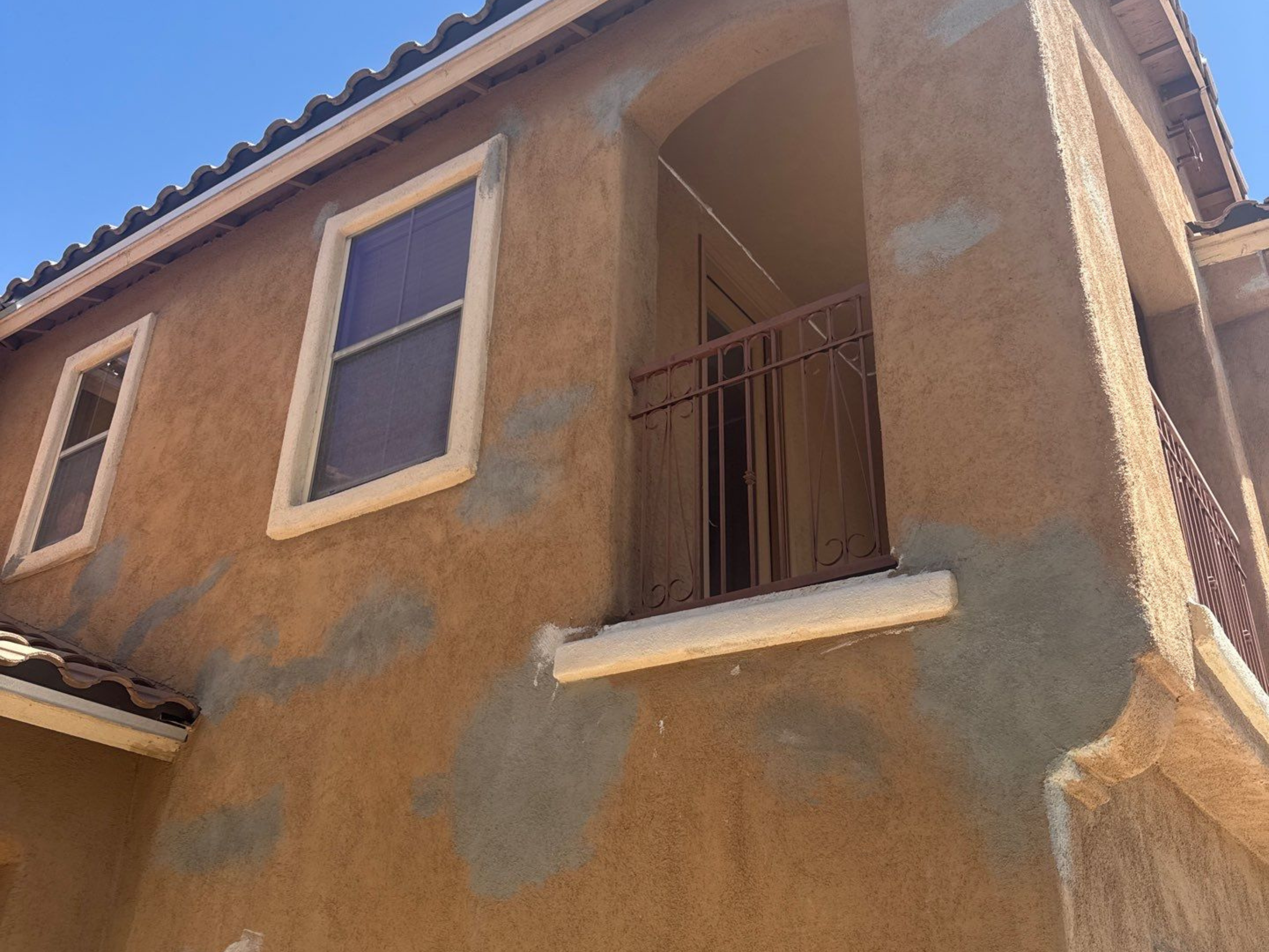 The image shows a beige stucco building with two windows and a small balcony with a metal railing under a clear blue sky.