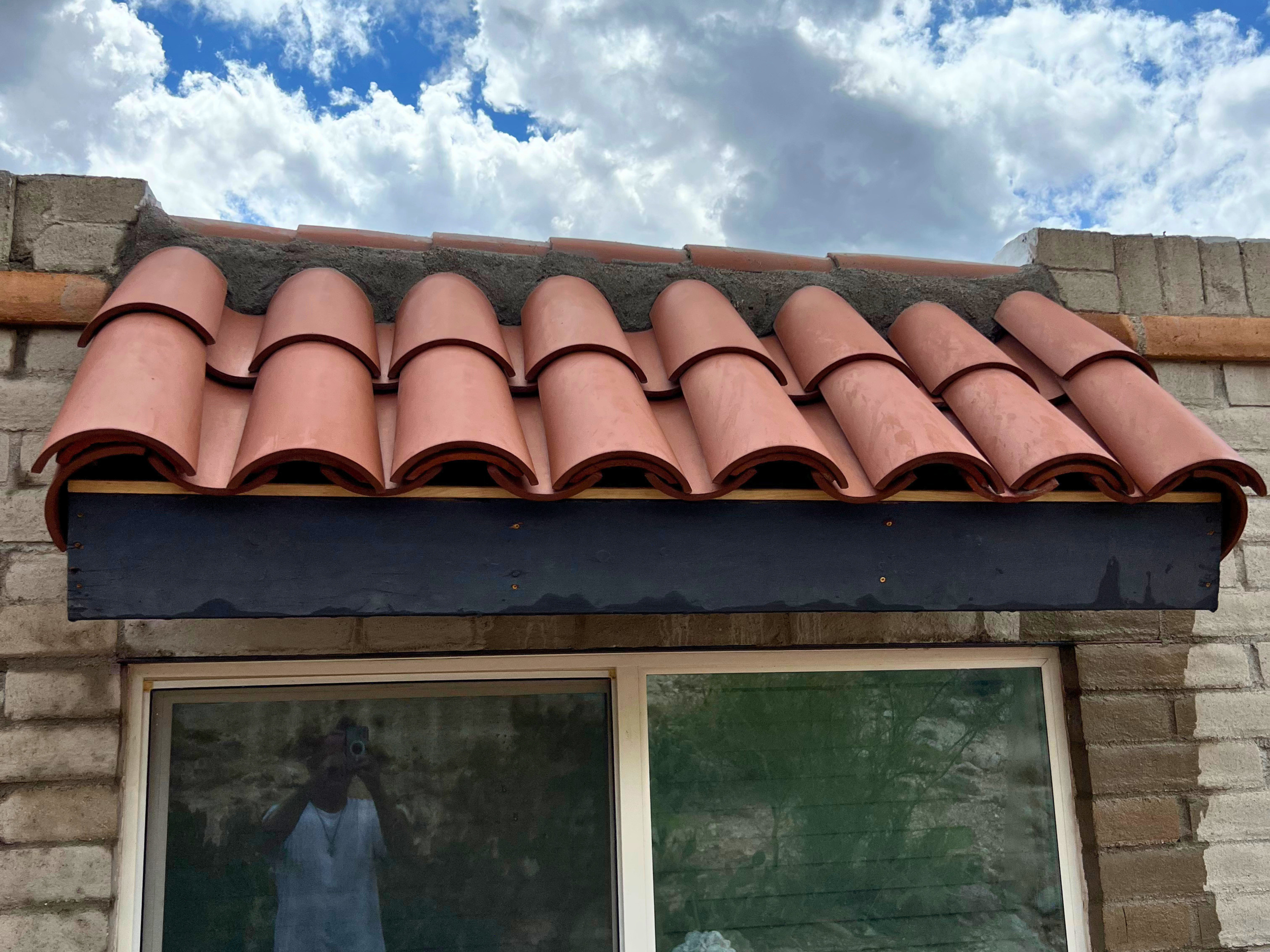 View of a brick house wall with a window and a decorative roof made of curved tiles, under a partly cloudy sky.