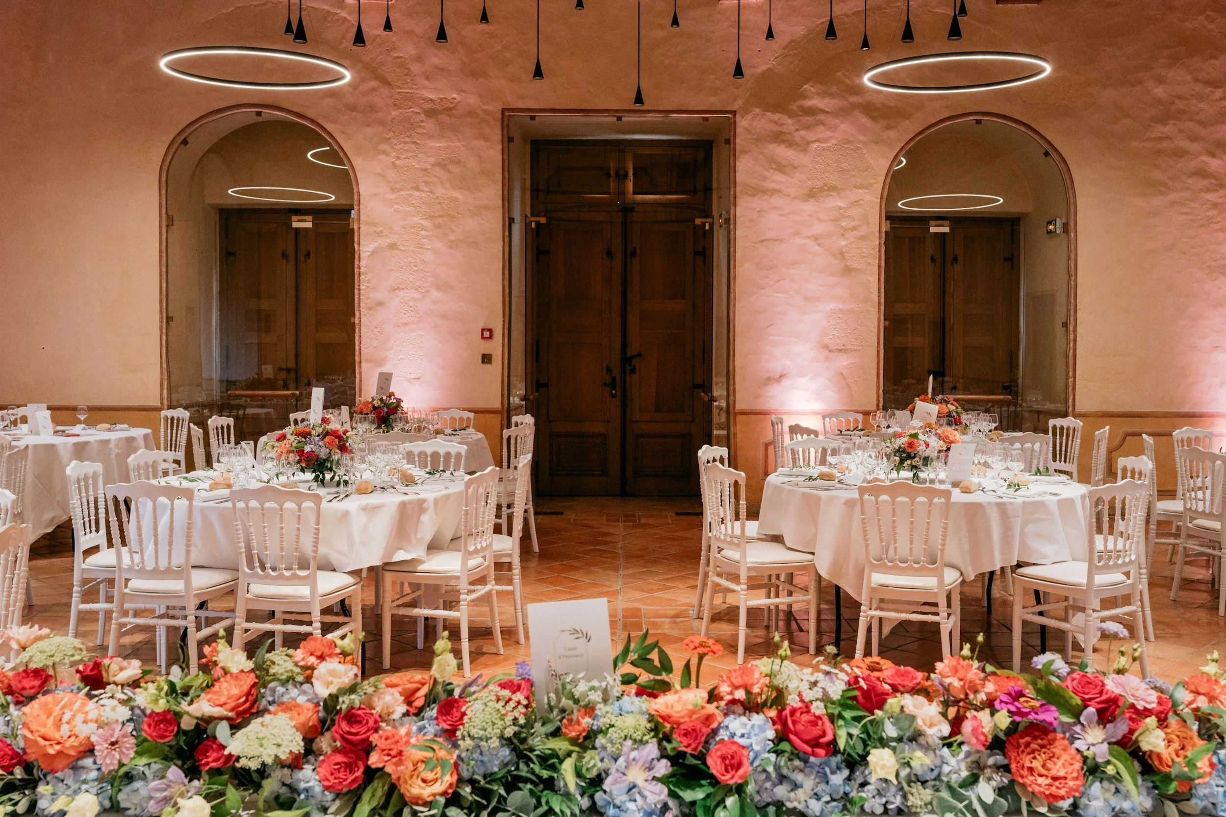 Elegant banquet hall with round tables set with white linens, wine glasses, and floral centerpieces. The room features arched windows, wooden doors, and soft pinkish lighting, with a large floral arrangement in the foreground.