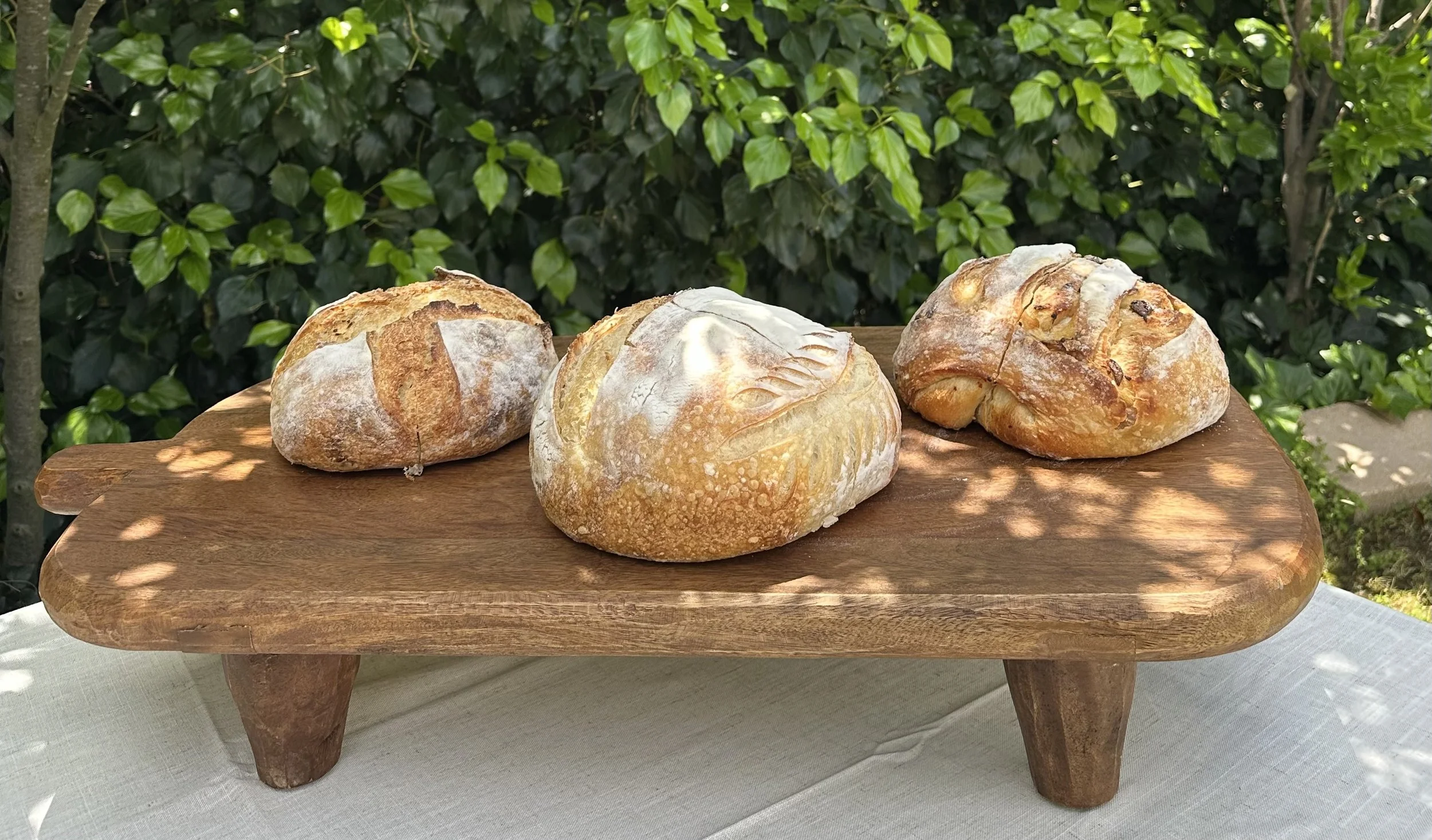 Three loaves of rustic bread on a wooden stand outside surrounded by green foliage.