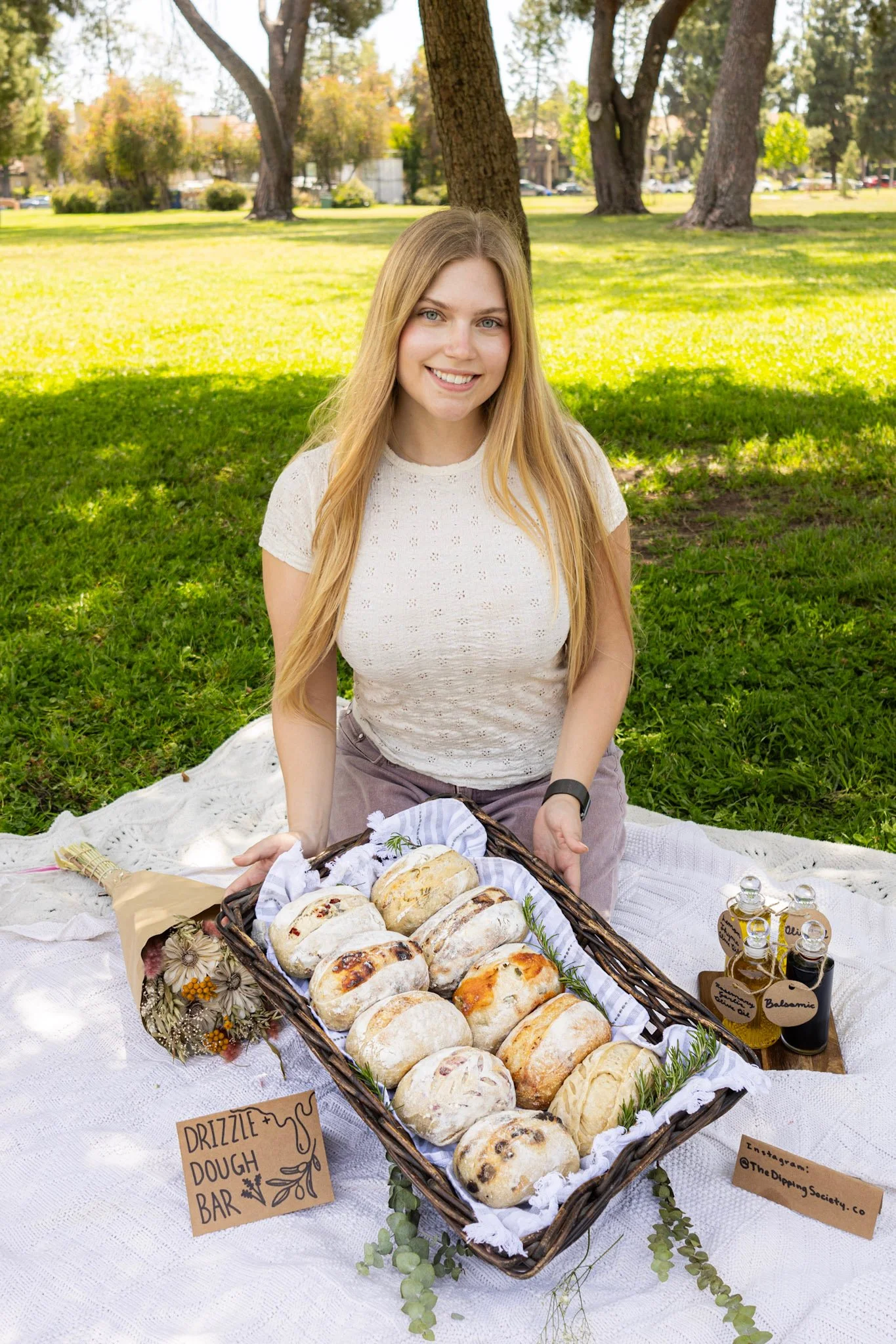 A woman with long blonde hair sitting on a blanket in a park during daytime, smiling, with a basket of assorted baked breads and signs indicating a drizzle dough bar and infused oil bottles set on the blanket.