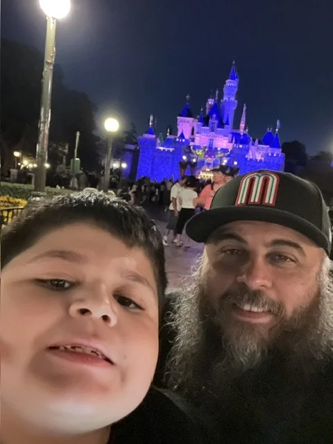 A boy with Down syndrome and a man taking a selfie in front of a lit-up castle at night at a Disney theme park.