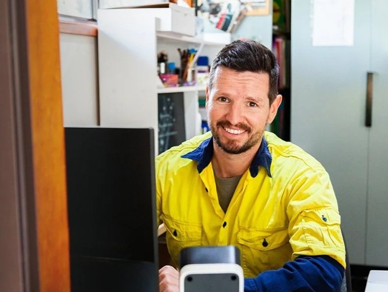 A man with dark hair and a beard, wearing a yellow jacket, smiling while sitting at a desk with a computer in an office or workspace.