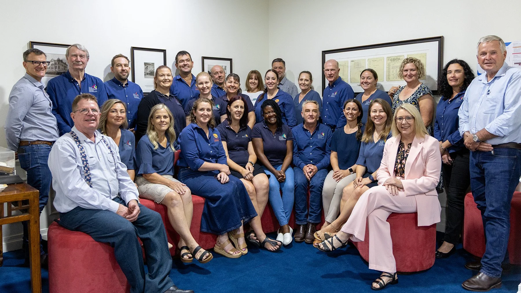 Group of diverse people in a room, seated and standing, posing for a group photo. Most are wearing blue shirts, with some in formal attire, in front of framed pictures on a white wall.
