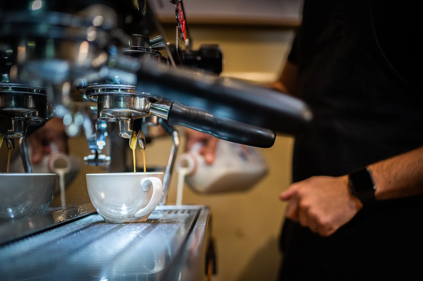 Close-up of a professional espresso machine dispensing coffee into white cups, with a barista preparing coffee in the background.