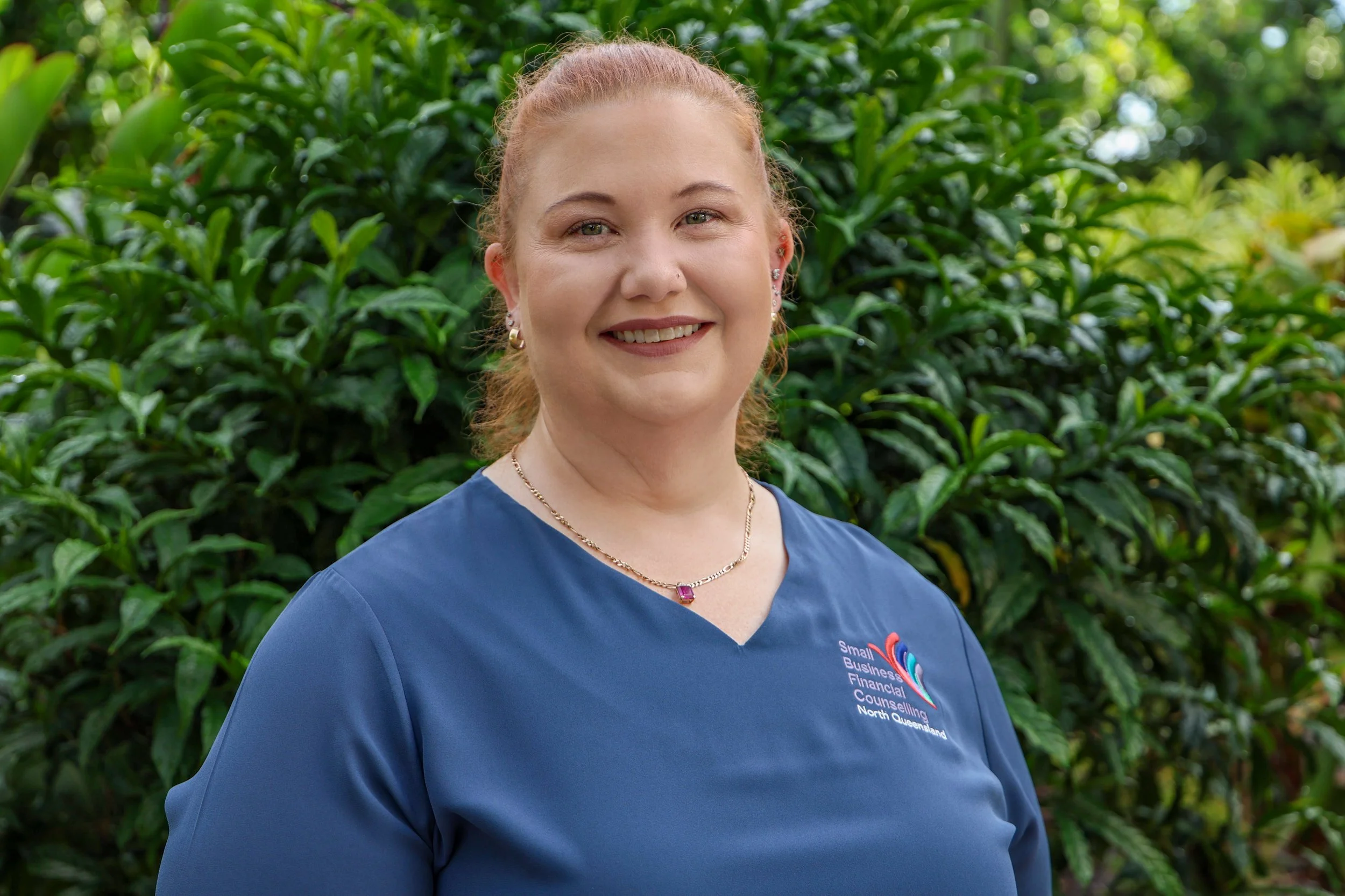 A woman with red hair tied back, wearing a blue shirt and jewelry, standing outdoors in front of green bushes.