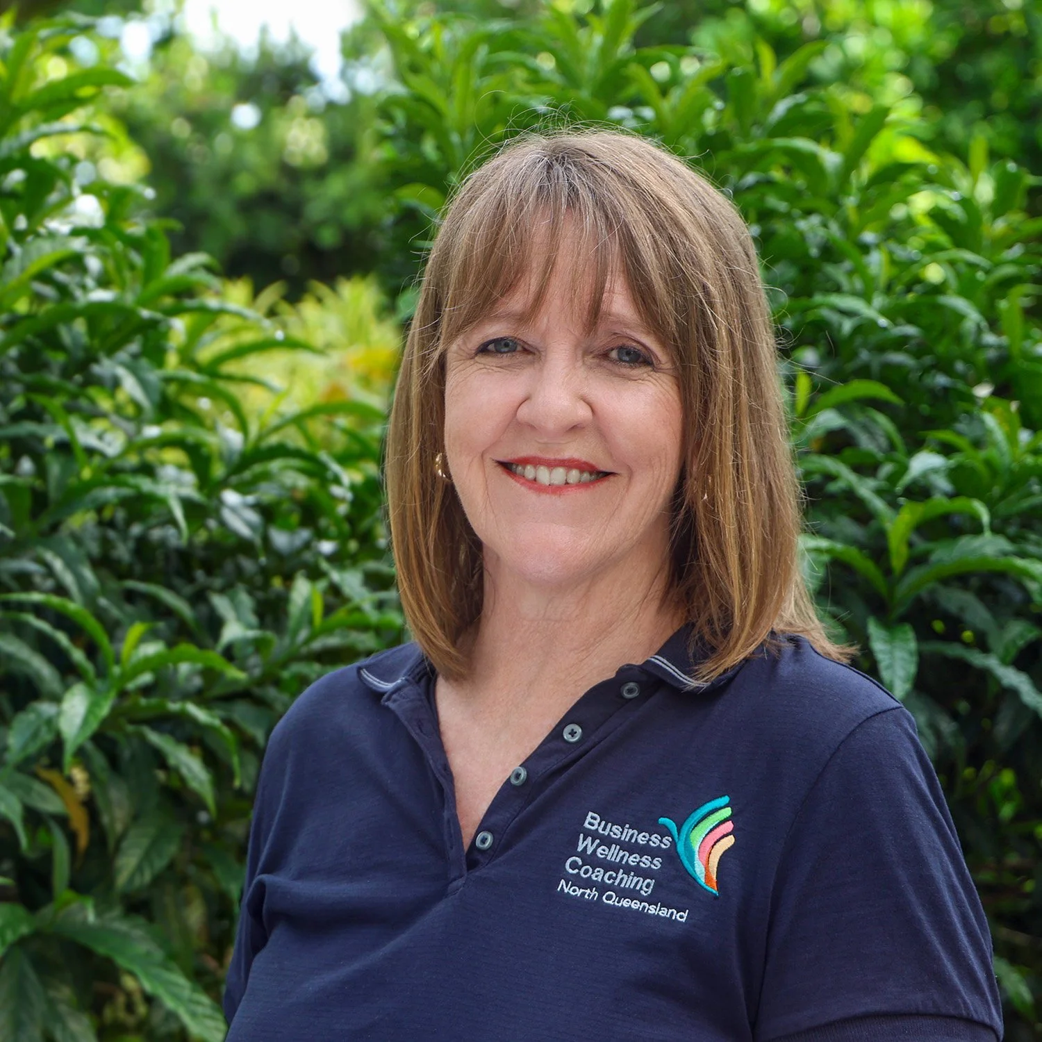 A smiling woman with shoulder-length light brown hair, wearing a navy blue polo shirt with a logo and text for Business Wellness Coaching North Queensland, standing outdoors with green leafy bushes in the background.