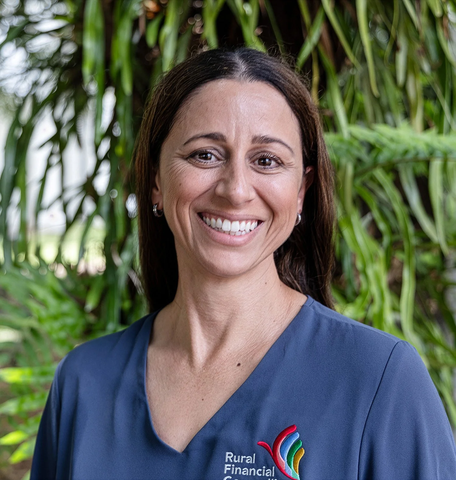 A woman with brown hair and a smile, wearing a navy blue shirt with a logo that says 'Rural Financial Corporation,' standing in front of green foliage.