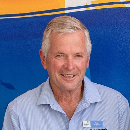 Smiling older man with gray hair wearing a light blue collared shirt and a name tag, standing in front of a blue background with yellow and navy stripes.