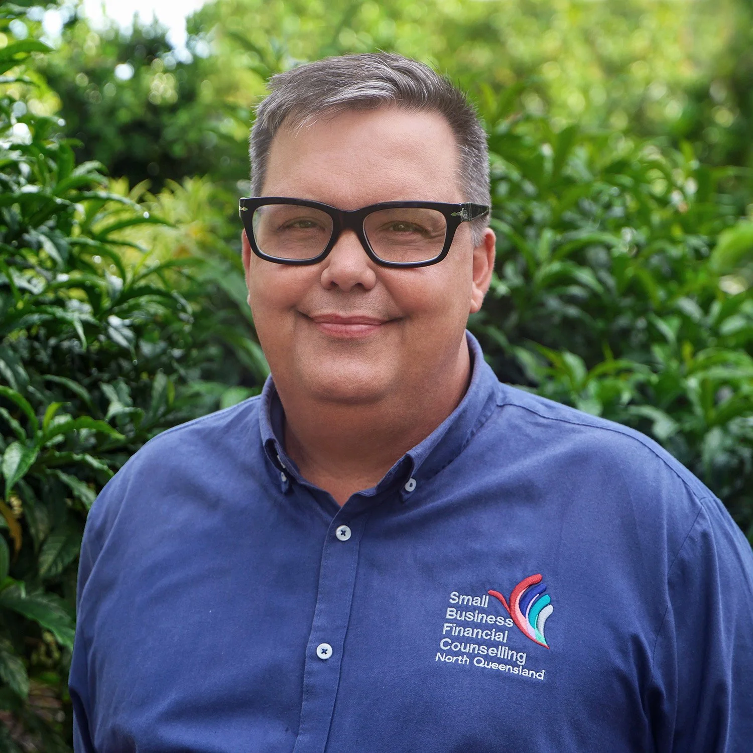 A middle-aged man with glasses and short gray hair, wearing a blue collared shirt with a logo that says 'Small Business Financial Counseling North Queensland,' standing outdoors in front of green foliage.