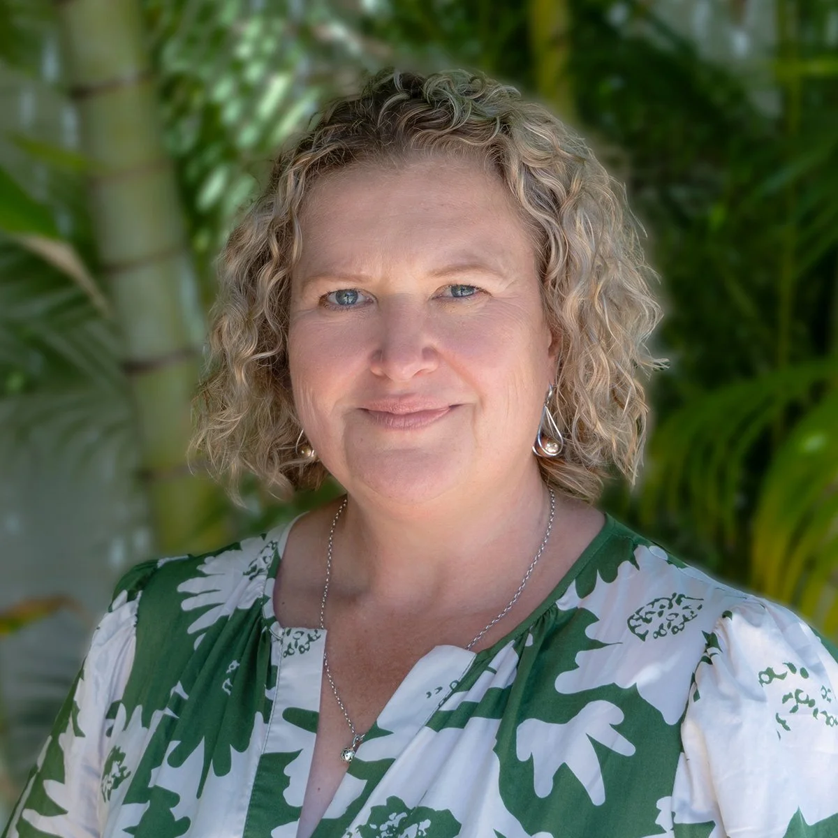 Close-up of a woman with curly blonde hair, wearing a green and white patterned dress, necklace, and earrings, standing outdoors with greenery in the background.