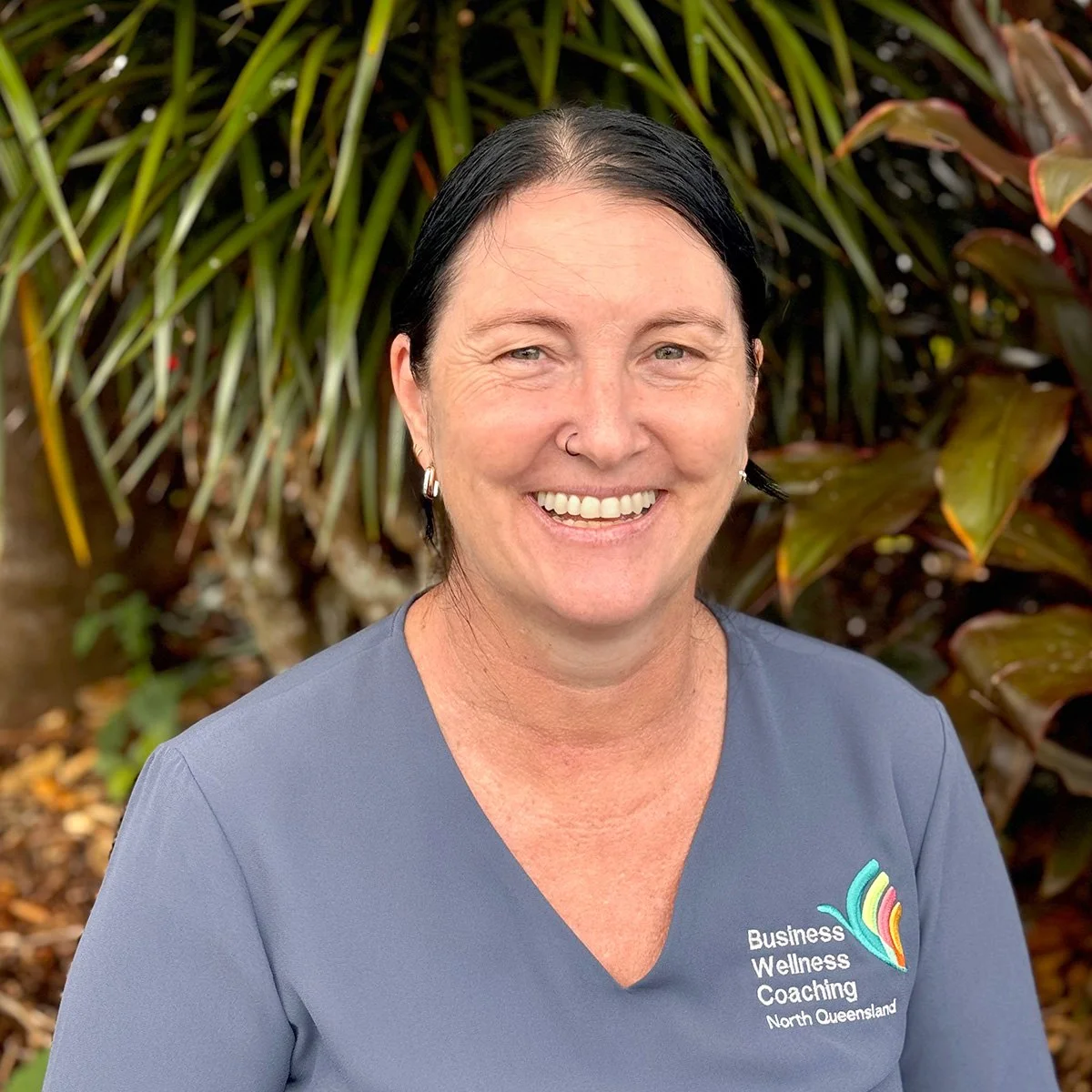 A woman smiling outdoors with green and brown tropical plants in the background. She has dark hair, is wearing earrings, a nose ring, and a gray V-neck shirt with a colorful logo and text that reads 'Business Wellness Coaching North Queensland.'