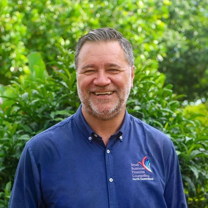 A smiling man wearing a blue collared shirt with a logo, standing outside in front of green foliage.