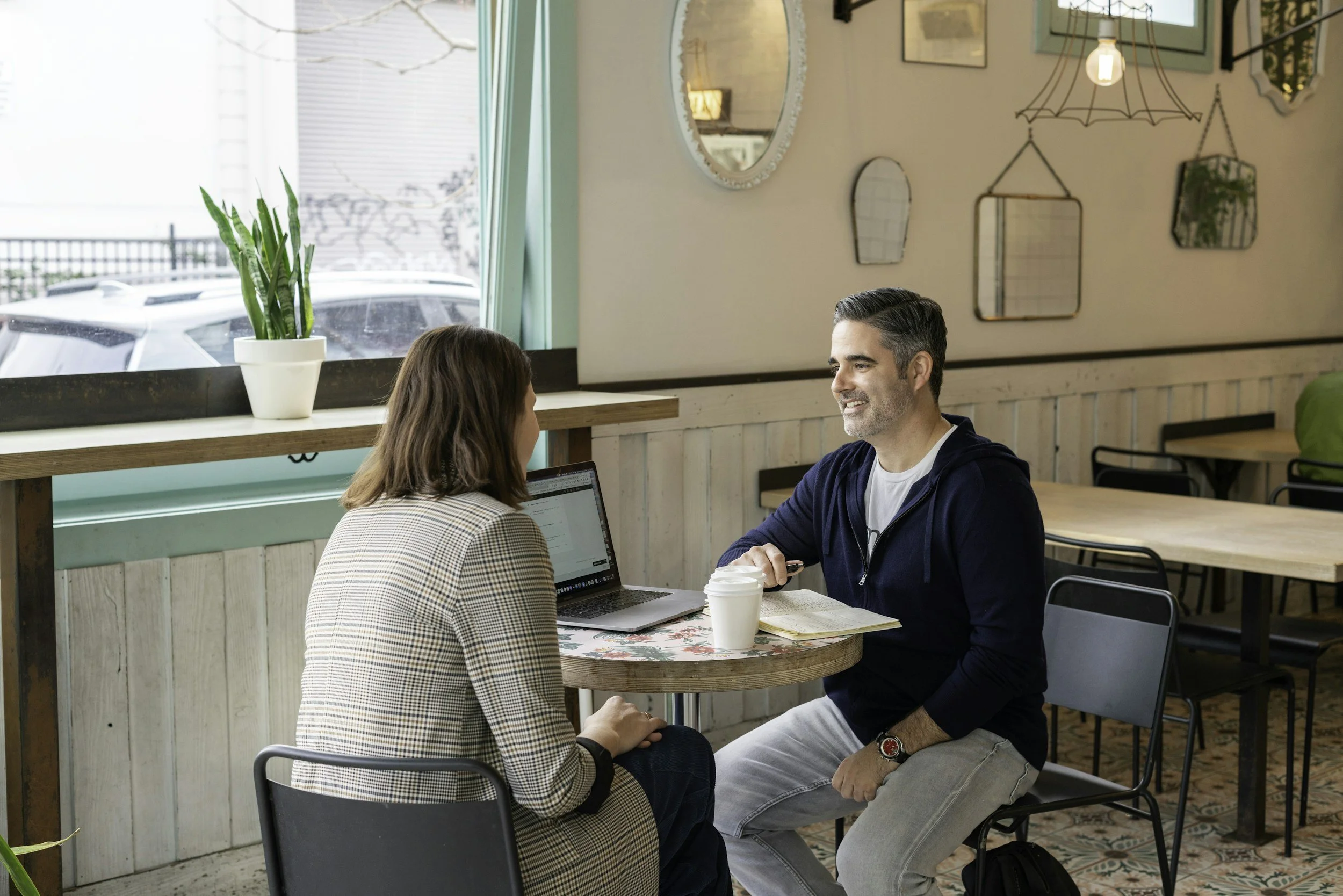 Two people, a woman and a man, are sitting at a table in a cafe, having a conversation. The woman is facing away from the camera, and the man is smiling and looking at her. The table has two cups of coffee, a laptop, and some papers. The cafe has a large window with a potted plant on the ledge, and decorative mirrors and artwork are hanging on the wall behind them.