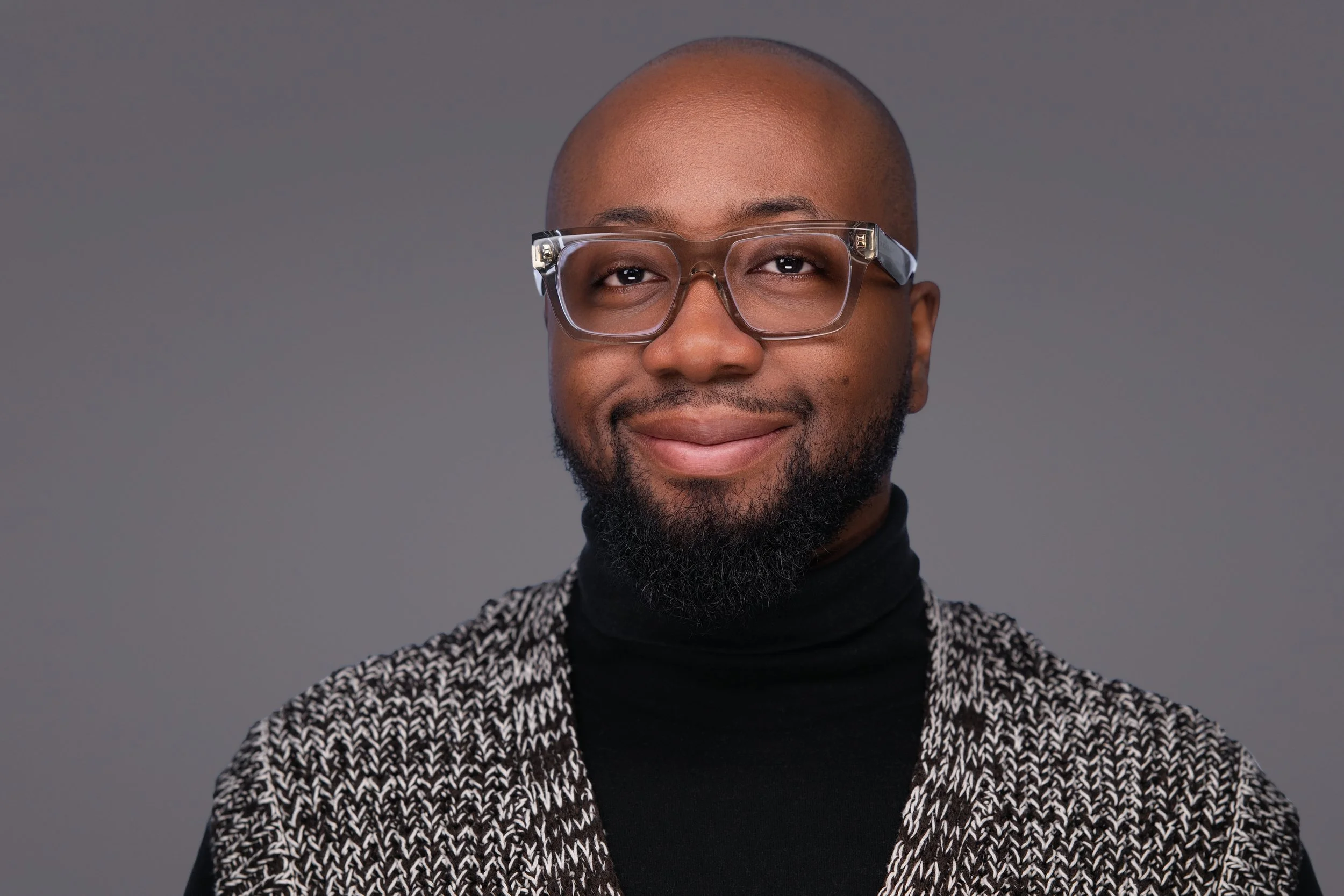 Portrait of a smiling African American man with a beard and bald head wearing large clear glasses, a black turtleneck, and a black-and-white patterned jacket against a neutral gray background.