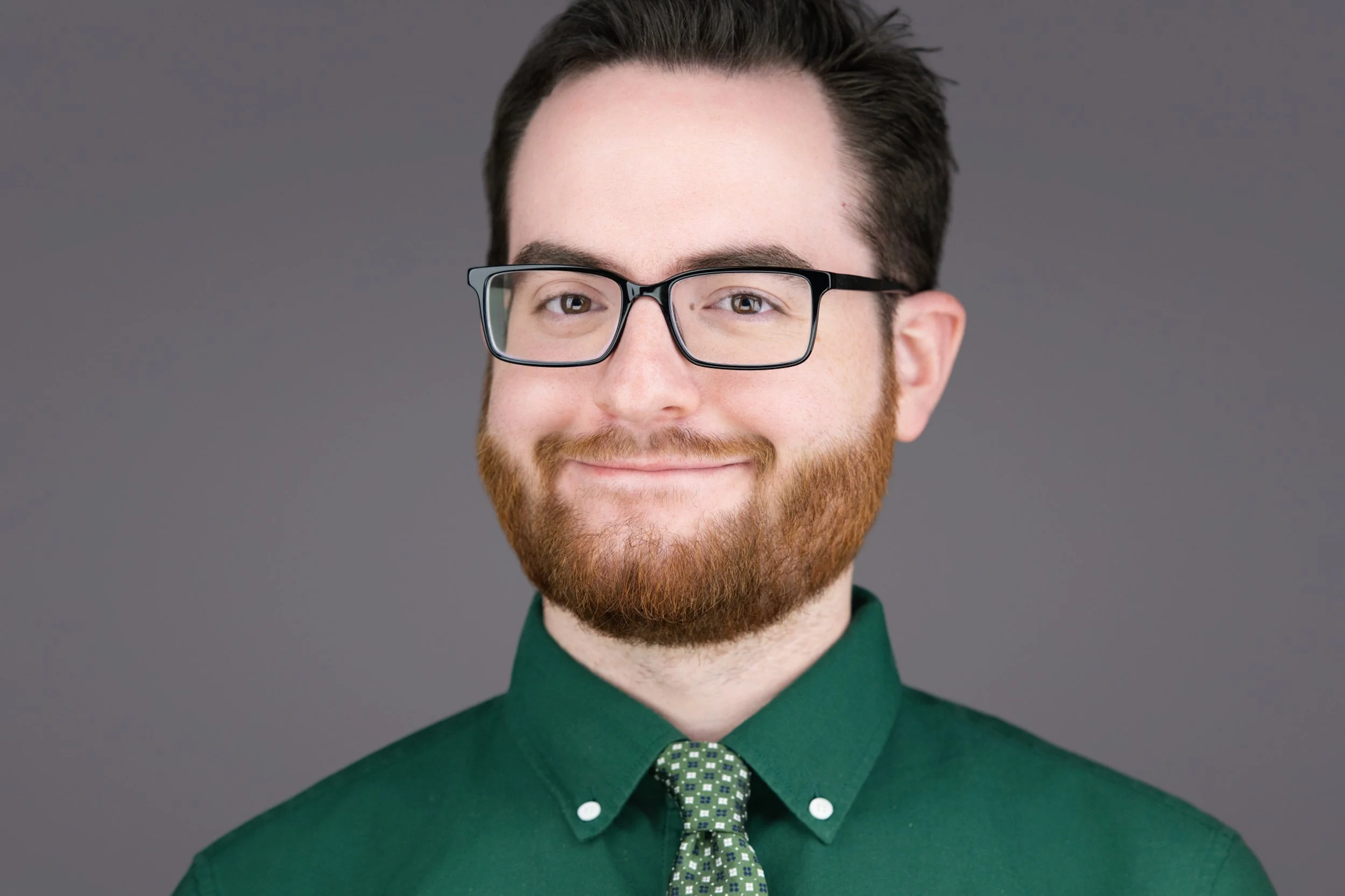 A portrait of a man with glasses, a beard, and a mustache, wearing a dark green shirt and a patterned tie, smiling against a gray background.