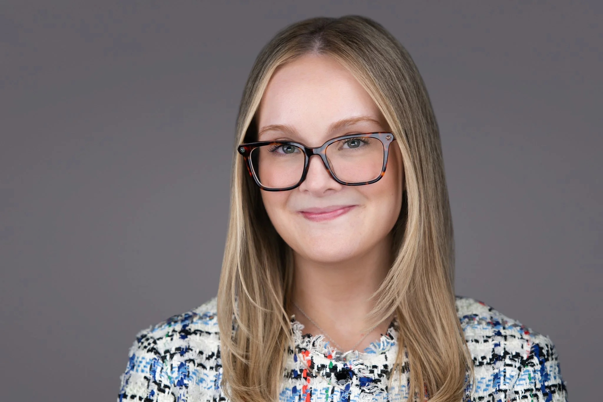 A woman with blonde hair wearing large, tortoiseshell glasses and a multicolored textured sweater smiling against a gray background.