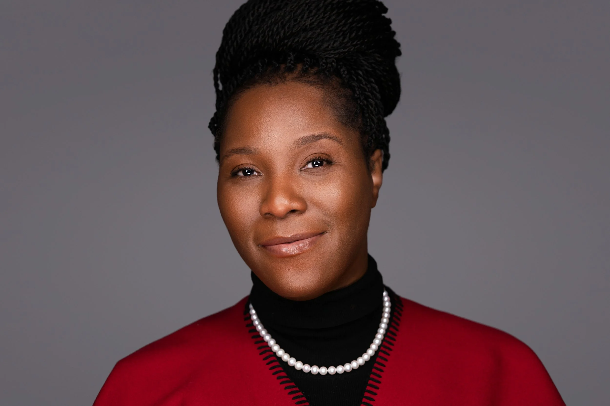 Portrait of an African American woman with styled braids, wearing a red blazer, black turtleneck, pearl necklace, and smiling against a gray background.