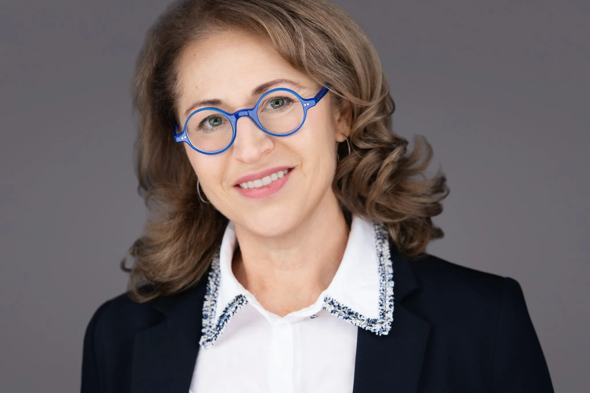 A woman with wavy light brown hair wearing blue glasses, a white shirt, and a dark blazer smiling against a gray background.