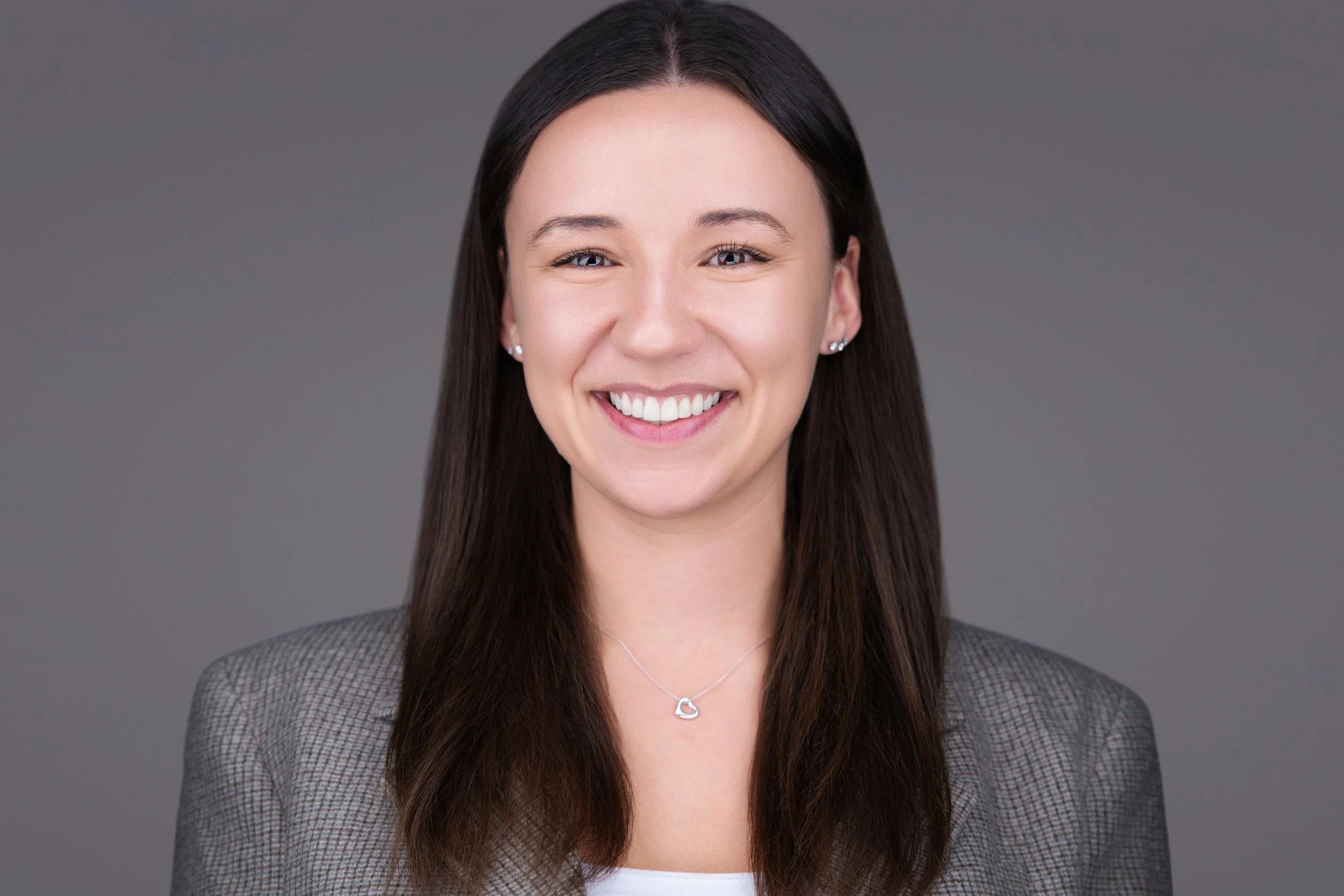 A young woman with long dark hair, smiling at the camera, wearing earrings, a necklace with a heart pendant, and a blazer, against a gray background.
