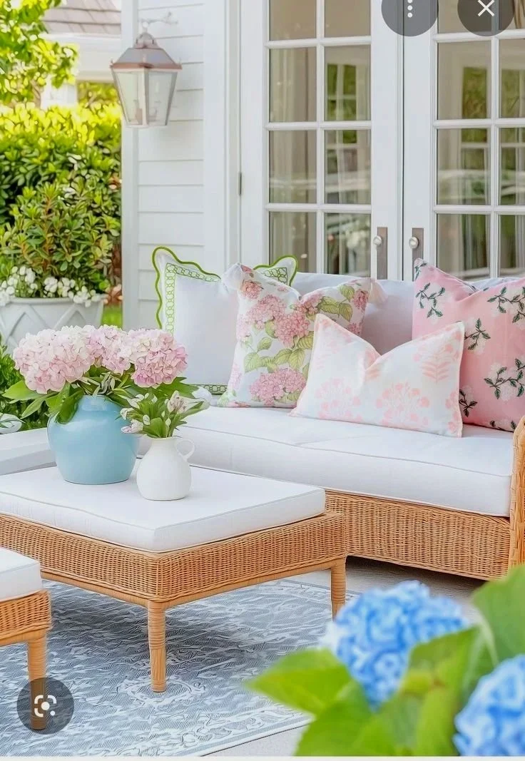 A cozy outdoor patio sitting area featuring a wicker sofa with light-colored cushions and floral pillows, a matching wicker coffee table with a white cushion top, a blue vase with pink hydrangeas, a small white pitcher, a hanging lantern, and French 