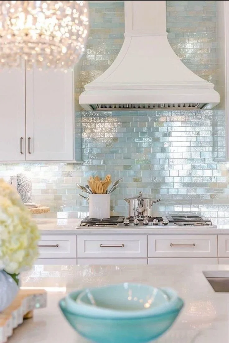 Modern kitchen with white cabinets, a white range hood, a blue backsplash, a pot with utensils on the stove, and decorative bowls and flowers in the foreground.