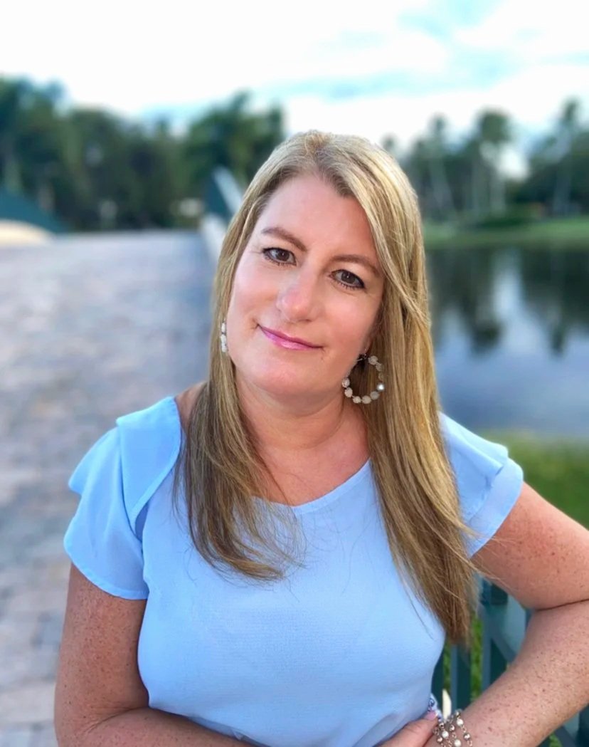 A woman with long light brown hair, wearing a light blue top and large hoop earrings, standing outdoors by a body of water with trees in the background