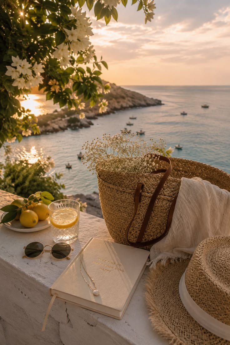 A seaside outdoor setting at sunset with a table featuring a book, sunglasses, a lemon drink, and a basket with flowers, overlooking water and boats.
