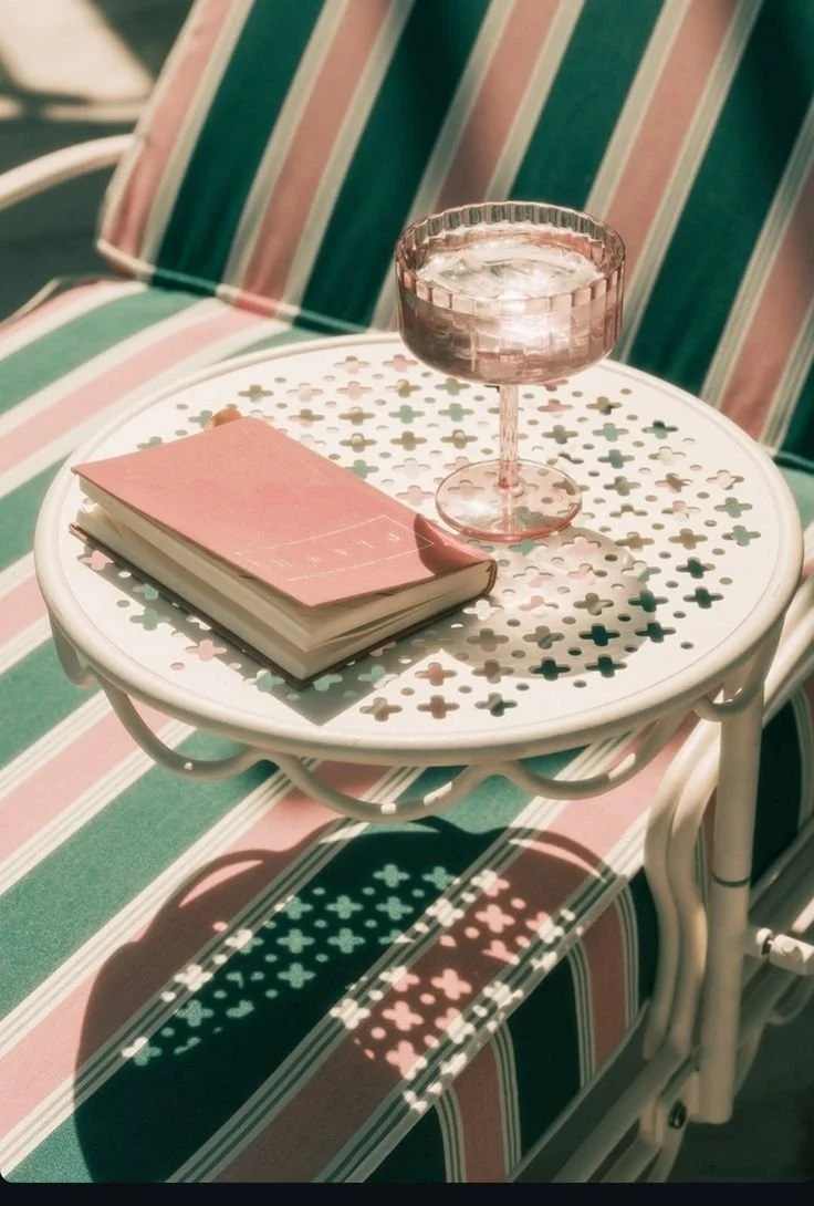 A pink book and a pink glass with a faceted design on top of a white round metal table with a decorative pattern, casting colorful shadows on a striped outdoor chair.