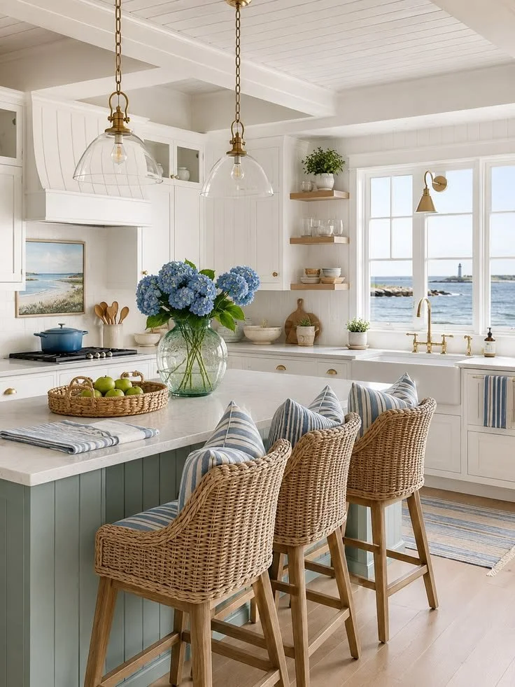 Bright coastal kitchen with white cabinets, a large window overlooking the ocean, blue hydrangea flowers in a glass vase, wicker bar stools with striped cushions, open shelves with dishes, and nautical decor.