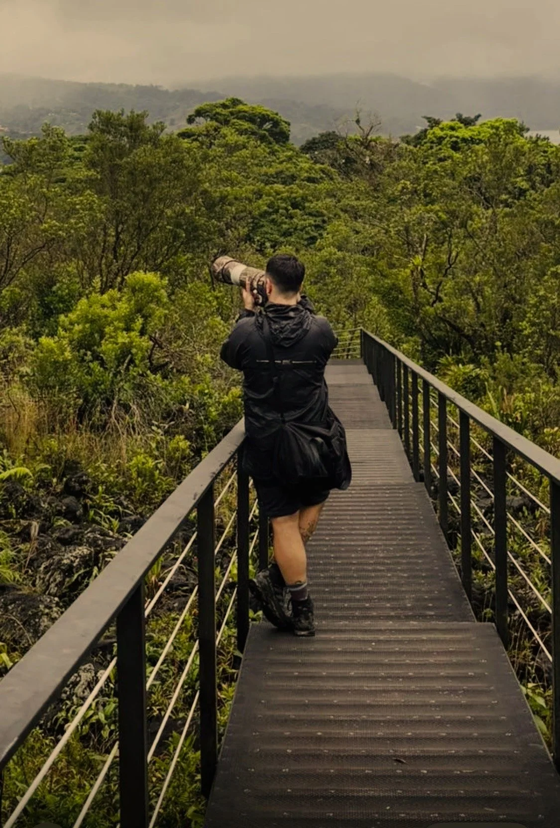 A person with short dark hair, wearing a black jacket, shorts, and hiking shoes, stands on a wooden bridge with metal railings, holding a camera with a large telephoto lens, surrounded by lush green forest, with mountains and cloudy sky in the background.