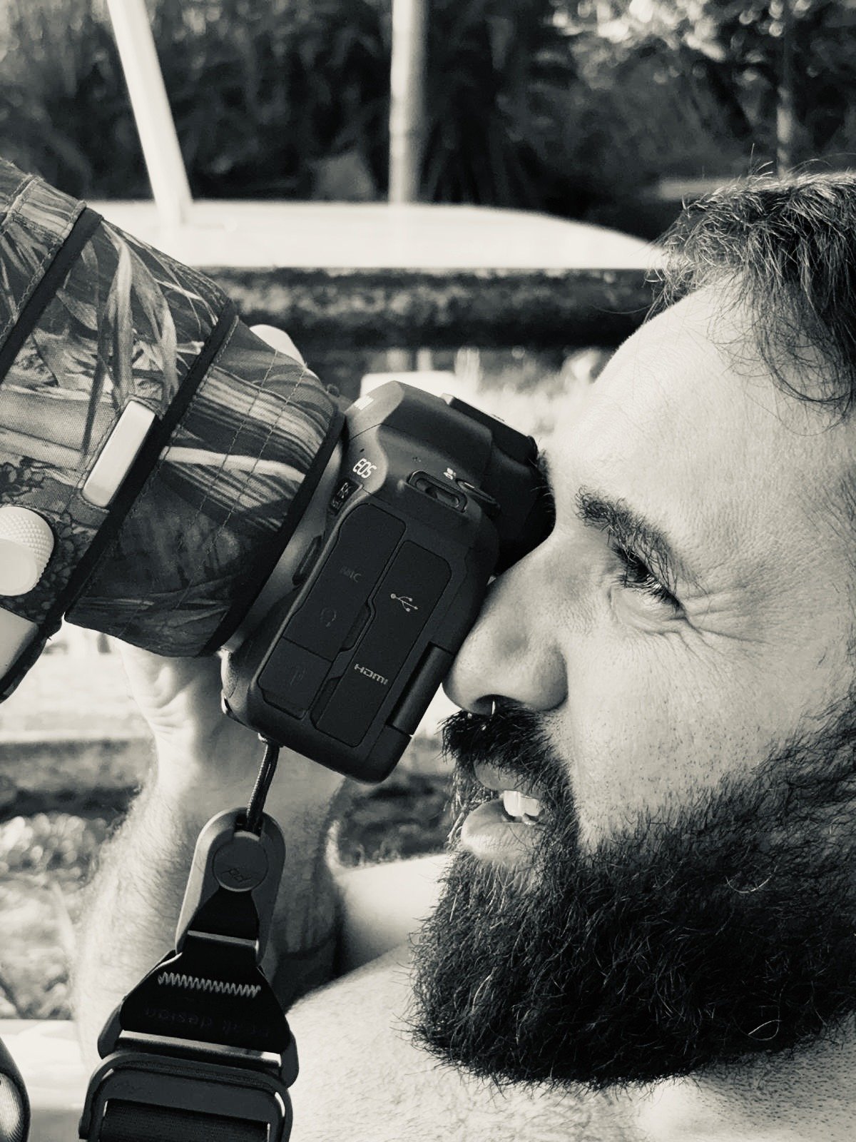 A man with a beard and mustache holding a camera against his face and looking through the viewfinder in an outdoor setting.