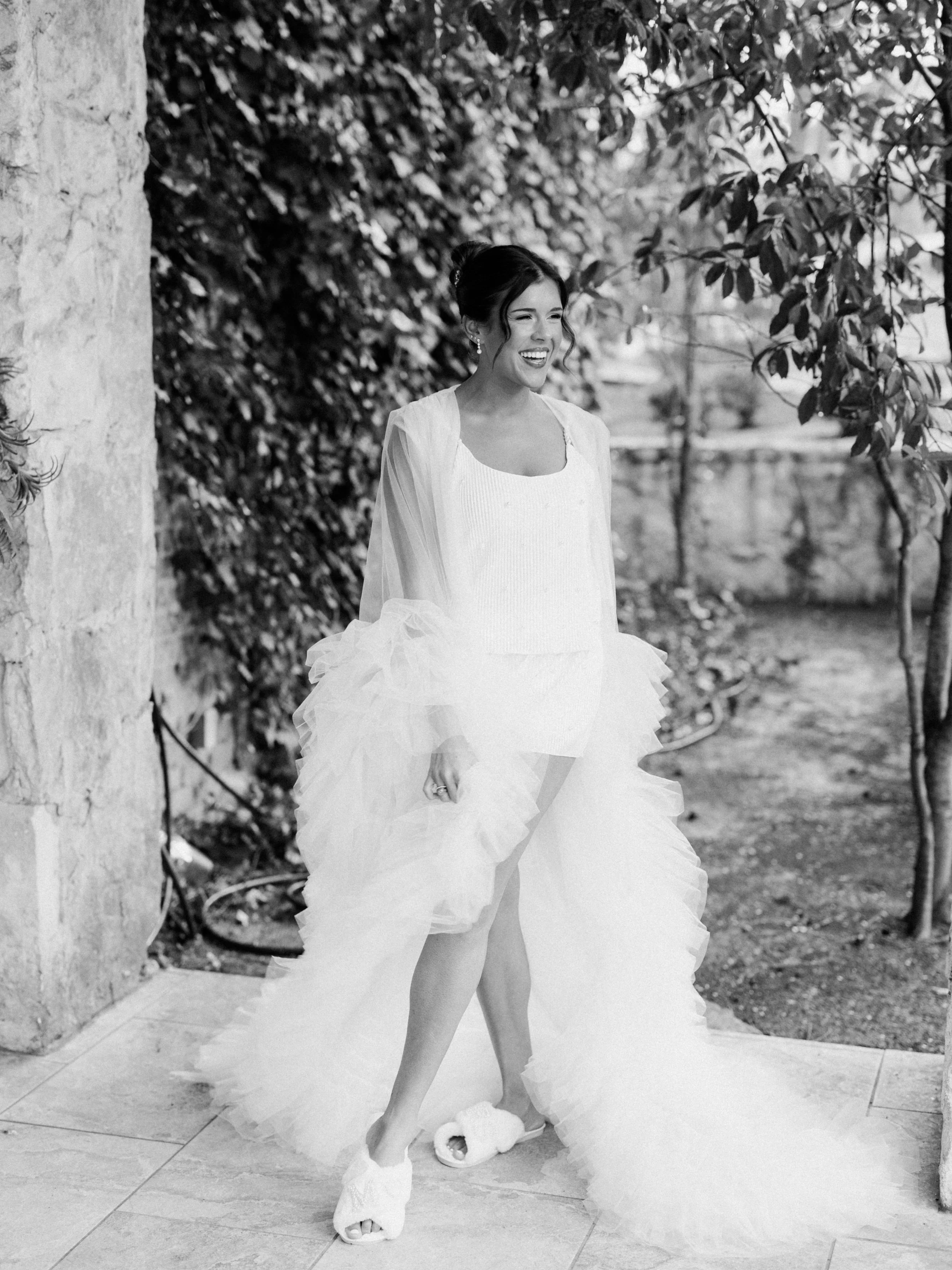 A woman in a wedding dress with a strapless bodice and puffed, layered skirt. She is standing outdoors in front of a vine-covered wall, smiling, with her hair styled up and wearing earrings.