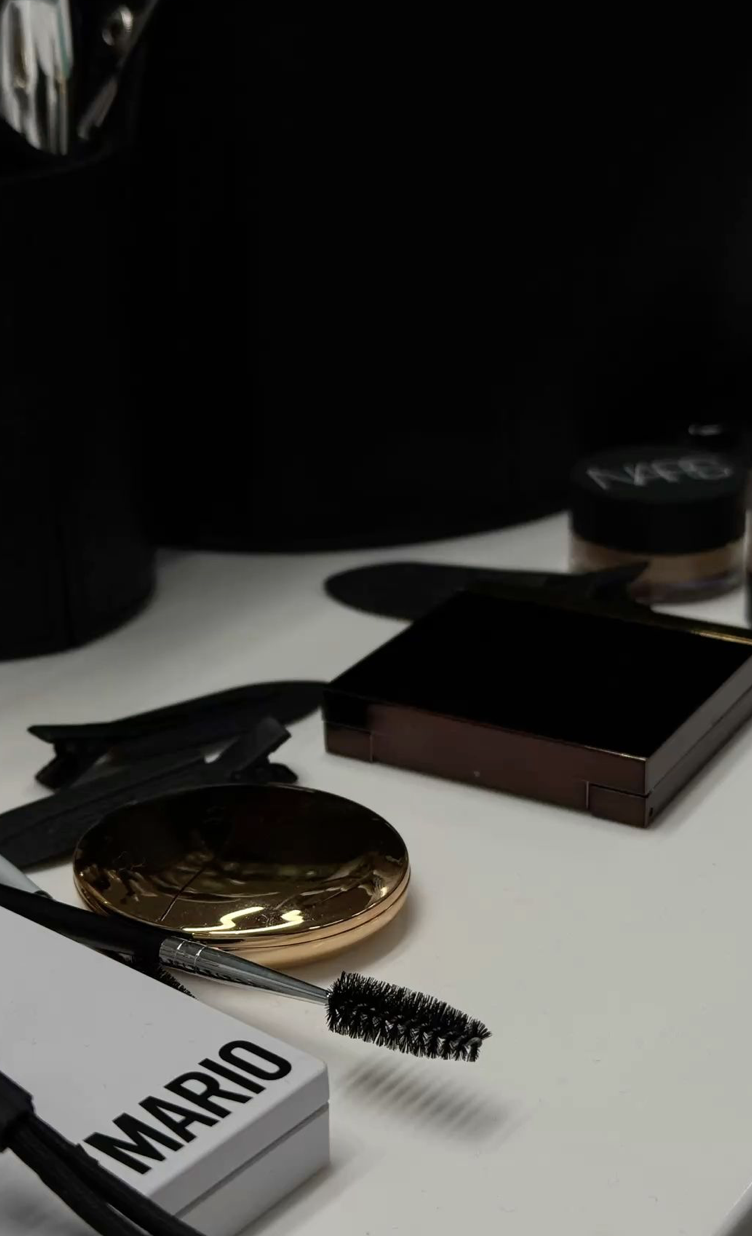Close-up of a makeup and cosmetic collection on a desk, including a gold compact mirror, mascara with a wand, a small square mirror, a black eyeshadow palette, and a container of loose powder, with a black desk organizer in the background.