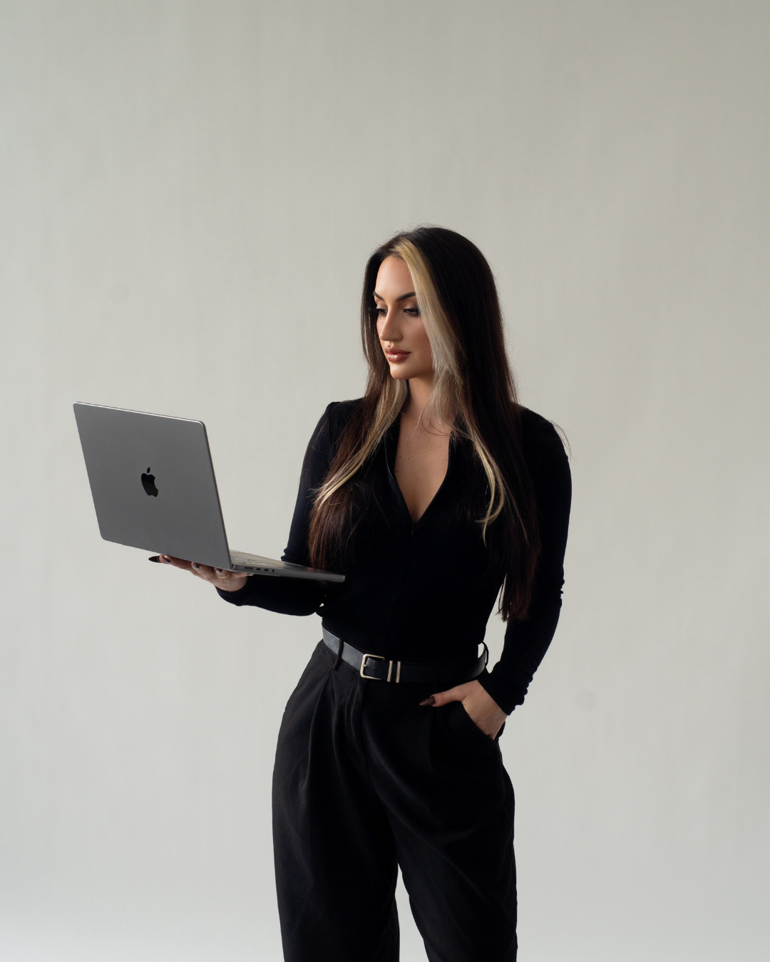 Woman with long dark hair and blonde highlights holding an open MacBook laptop, standing against a plain light background.