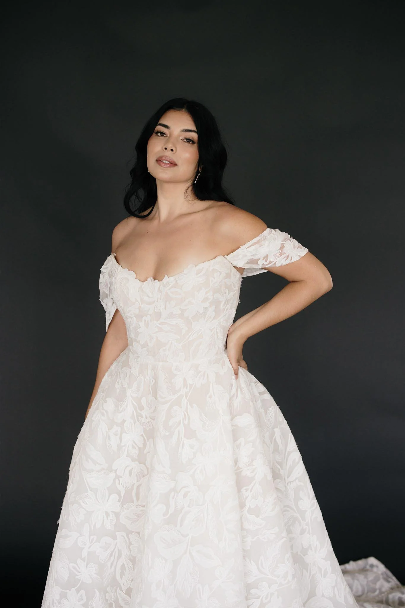 A woman with black hair and earrings wearing an off-the-shoulder white floral wedding dress standing against a dark background.