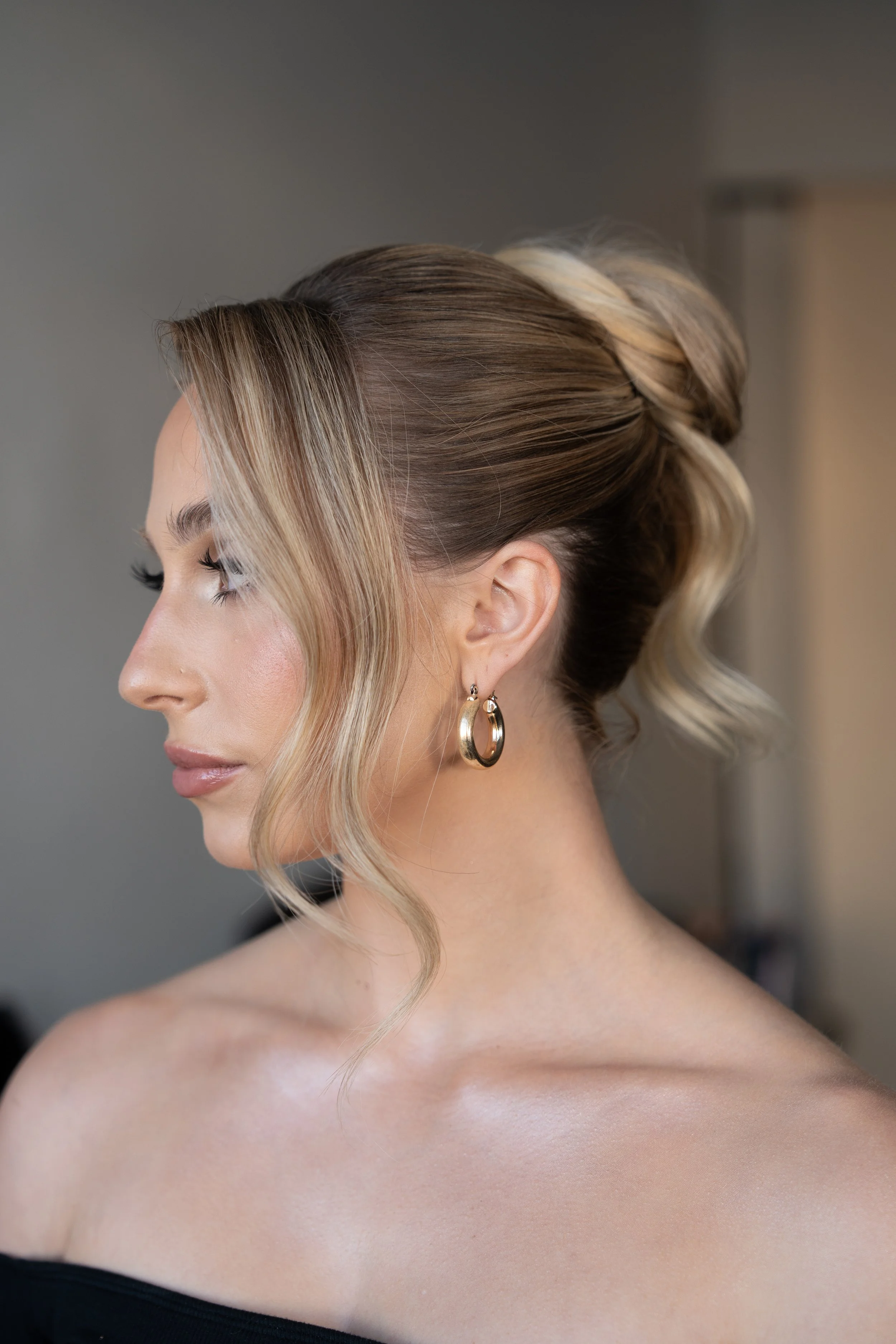 Side profile of a woman with styled blonde hair in soft waves, wearing gold hoop earrings, and a black off-the-shoulder top, with a neutral background.