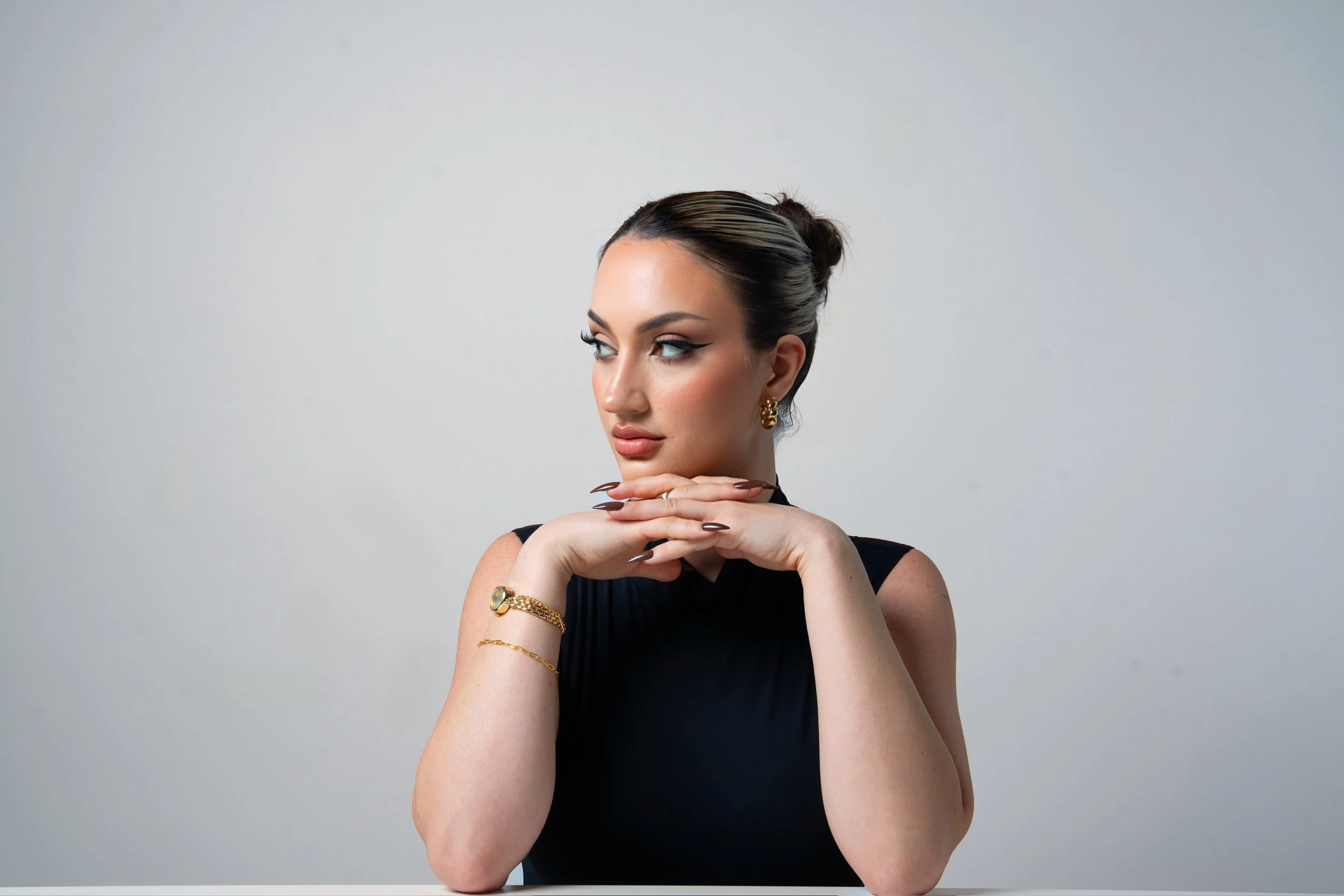 A woman with dark hair styled in an elegant updo, wearing makeup and gold jewelry, gazing to the side with her hands softly touching her chin, against a plain light gray background.