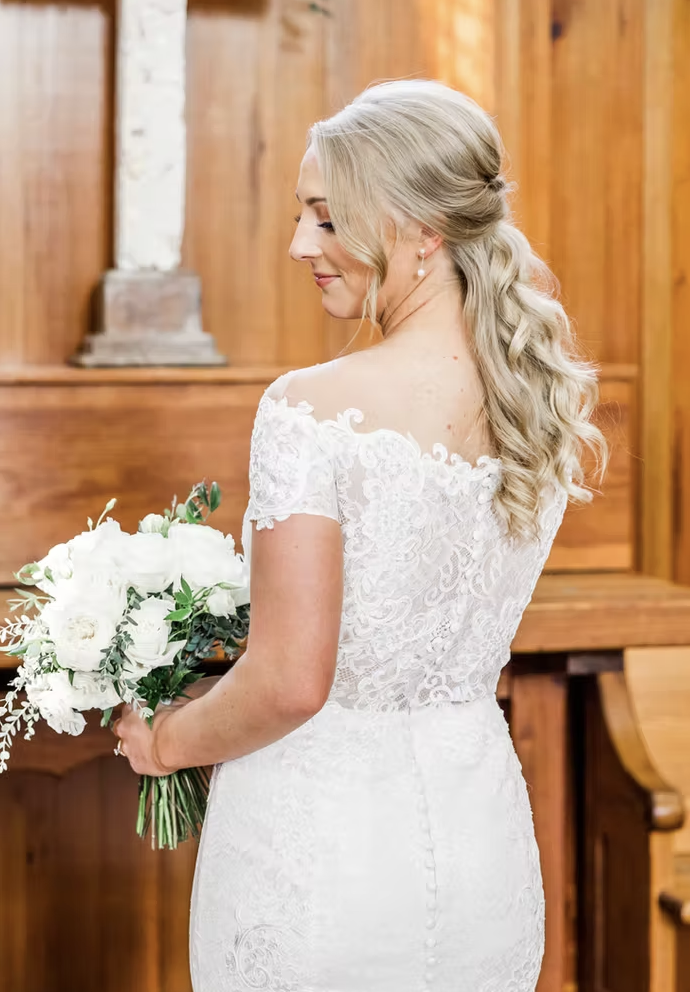 A bride with long, wavy blonde hair in a lace wedding gown holding a bouquet of white flowers.
