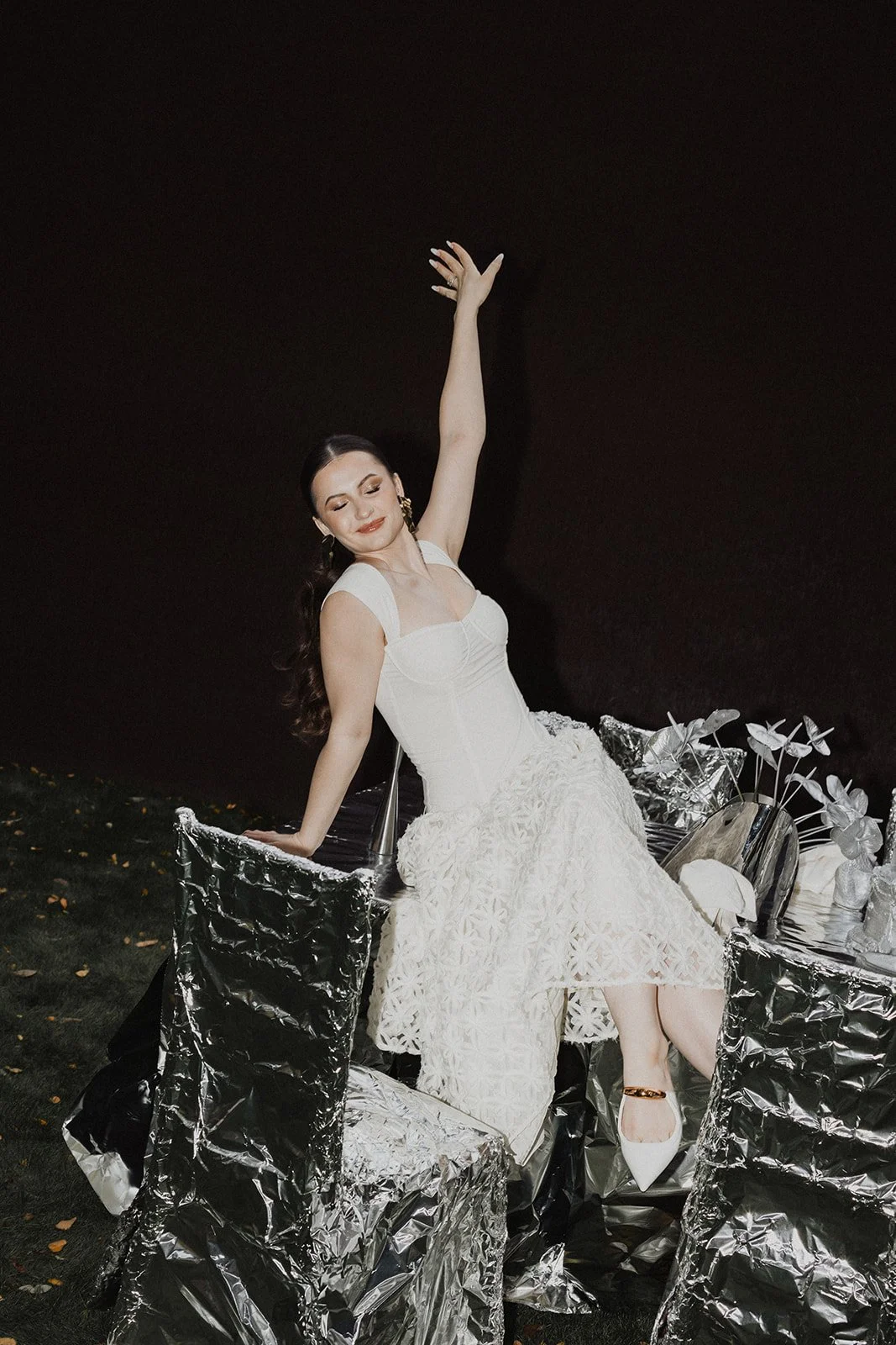 A woman in a white dress with a floral pattern, sitting on a silver foil-covered chair, raising her right arm, and smiling with closed eyes.